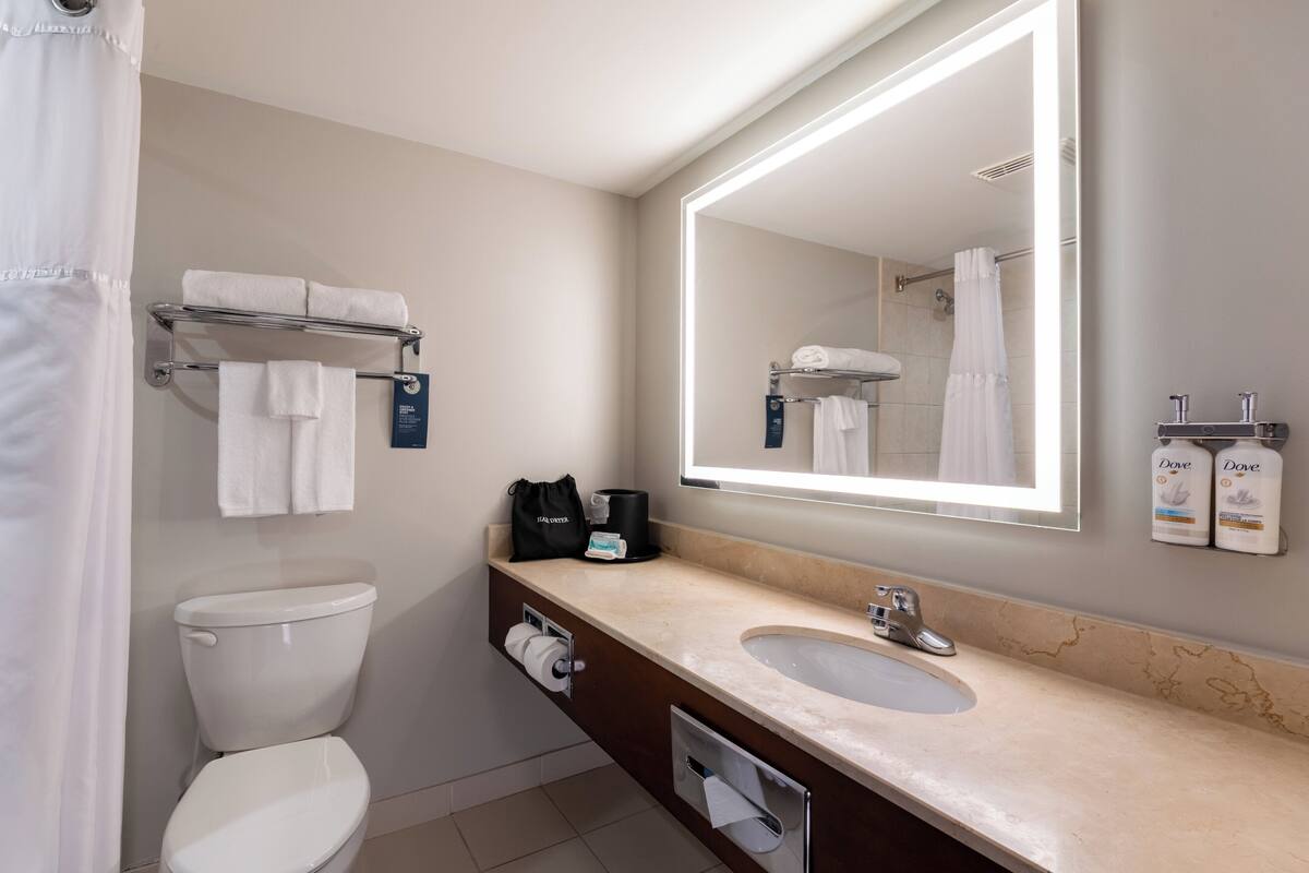 Un baño moderno con inodoro, lavabo y un espejo grande. Hay toallas en un perchero, una cafetera y artículos de aseo en la encimera.