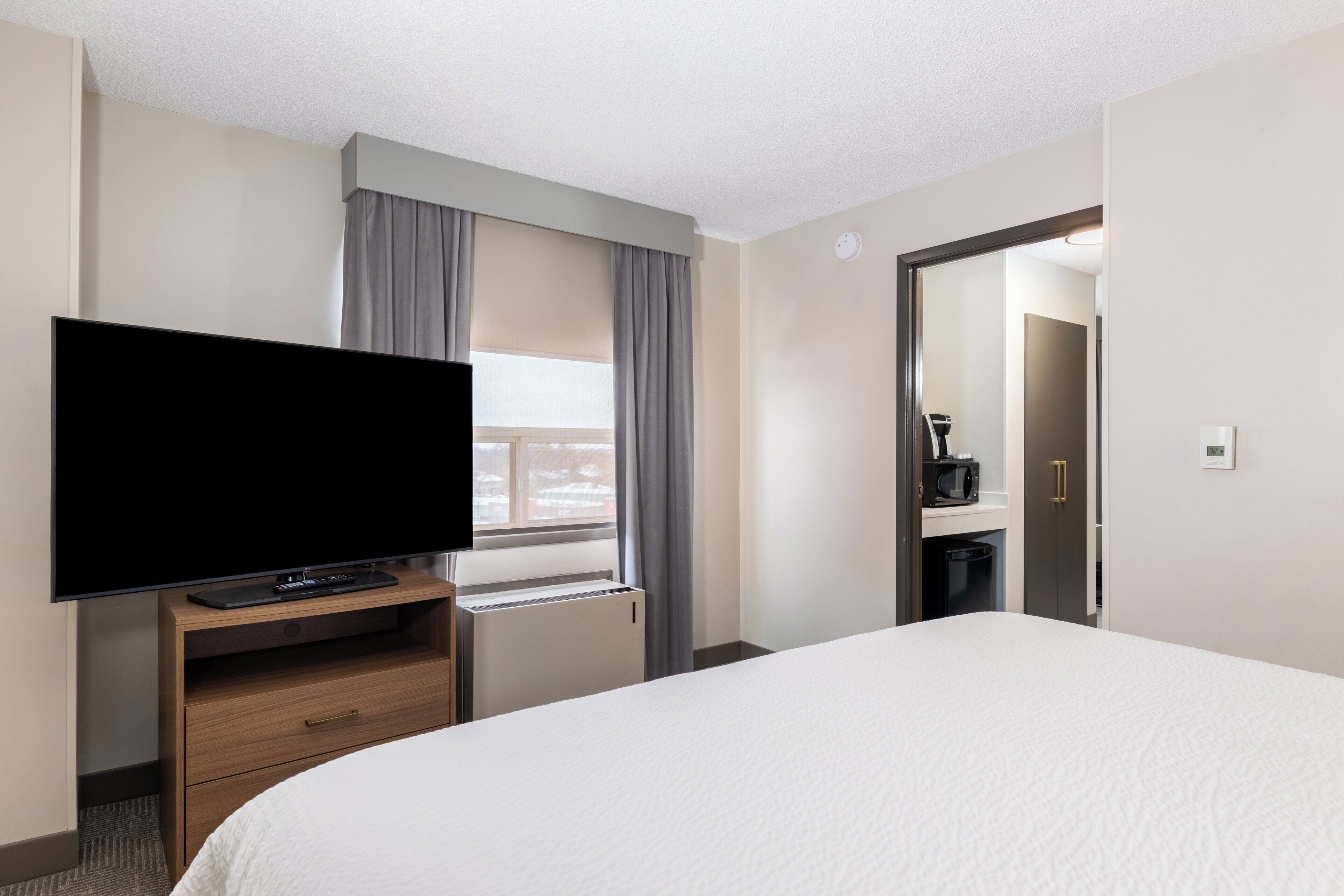 Una habitación de hotel con una cama grande, un televisor de pantalla plana sobre un soporte de madera, una ventana con cortinas y una puerta que da a un baño.