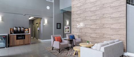 A spacious lobby with a modern design, featuring a seating area with sofas and armchairs, a coffee station, and a large wooden accent wall.