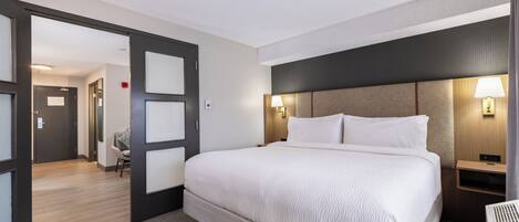 Habitación de hotel con una cama grande, un escritorio y una silla. Se ve una puerta que da a otra habitación y un pasillo.