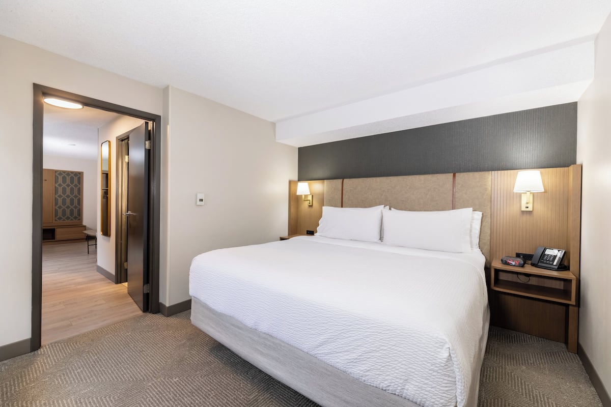 Una habitación de hotel con una cama grande, lámparas de noche, una mesita de noche y una puerta que da a un pasillo.