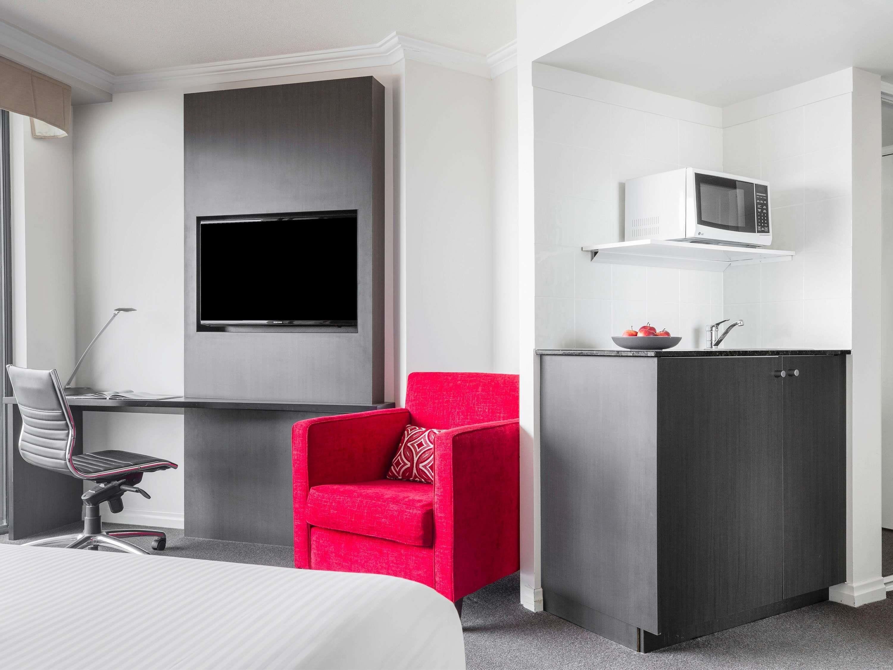 A hotel room with a bed, a desk with a computer, a TV, a microwave, and a red chair.