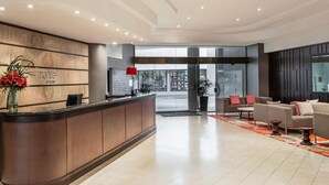 A hotel reception area with a wooden reception desk, a red lamp, and a seating area with sofas and tables. - Mantra on Queen (Brisbane)