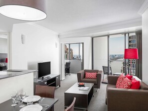 A modern hotel room with a dining area, a TV, a sofa, and a view of the city.