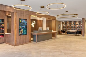 Un área de recepción con un mostrador de madera, un letrero iluminado con la letra 'H' y una zona de estar con un muro estampado y un sofá.