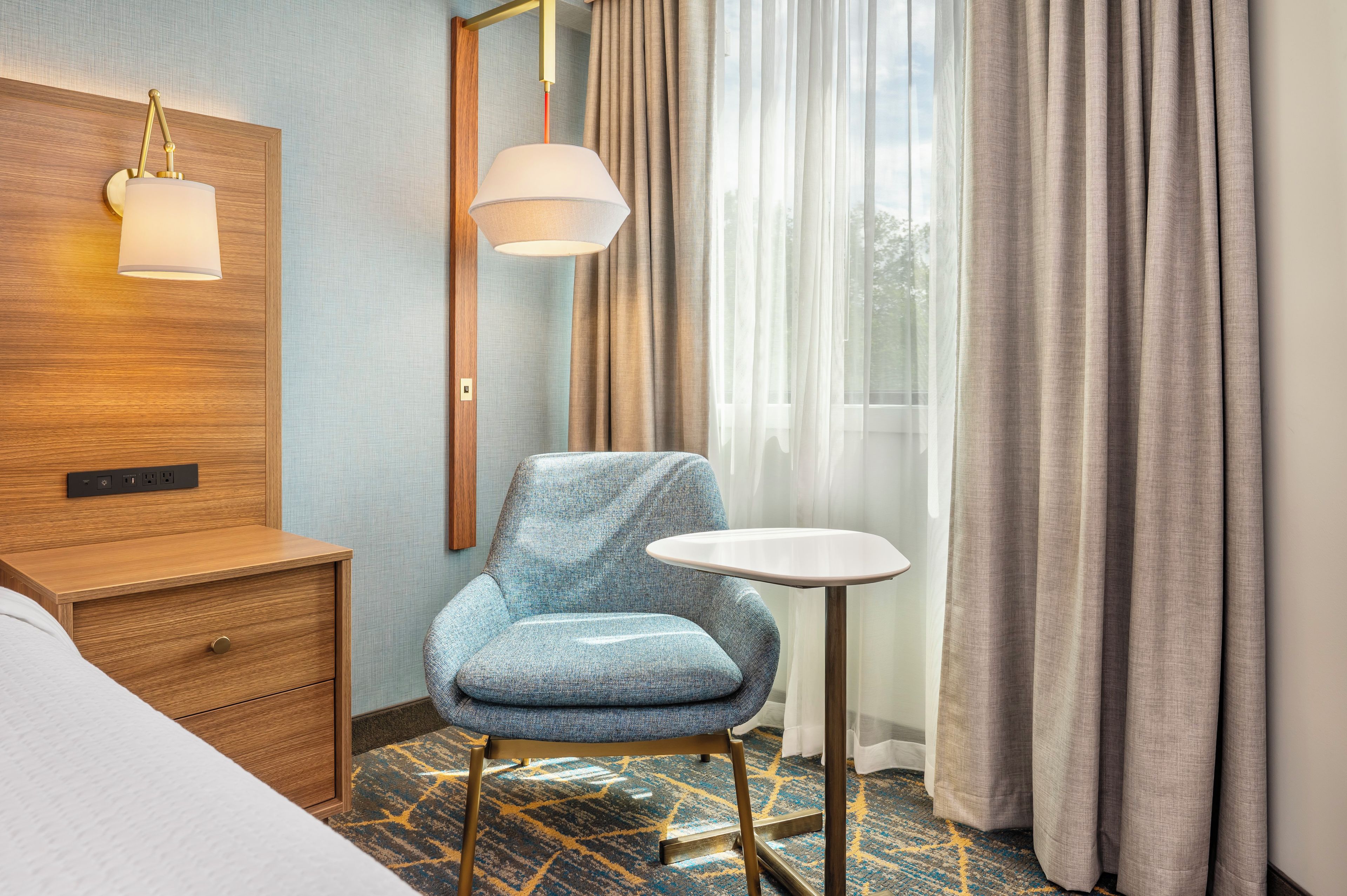 A hotel room with a bed, a wooden nightstand, a blue upholstered chair, a small white table, a wall-mounted lamp, and a window with sheer curtains.