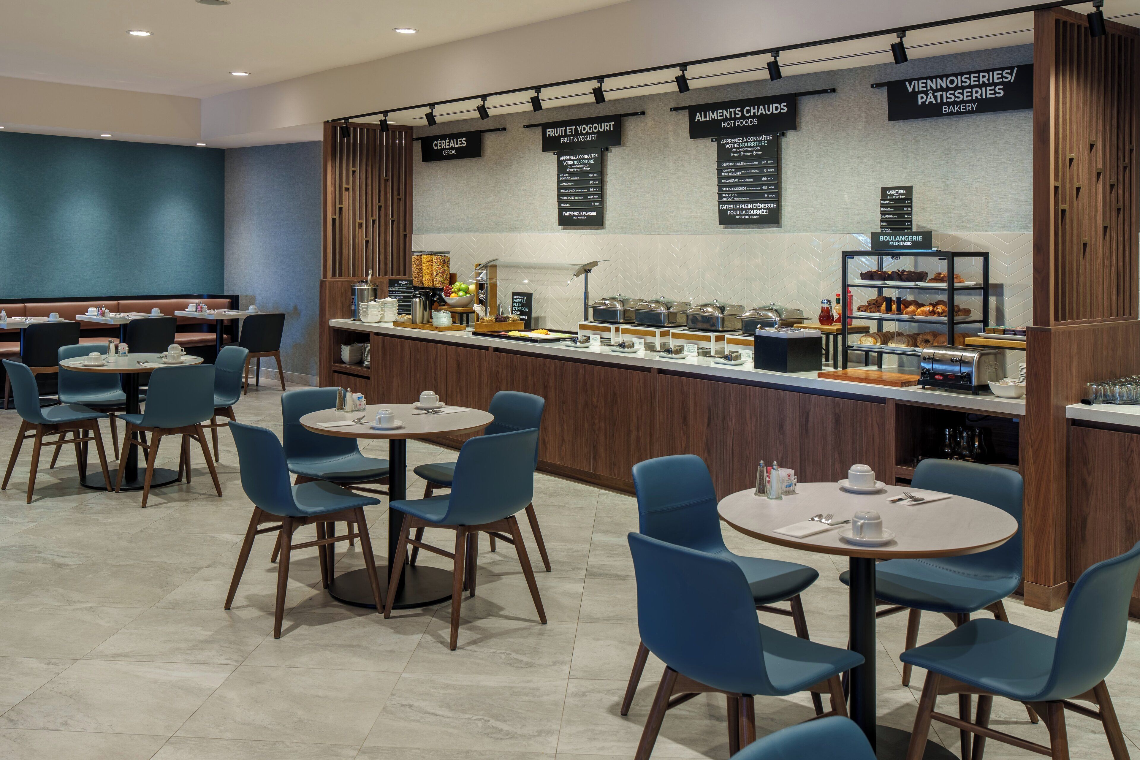 A modern café with round tables and blue chairs, a counter with various food items, and menu boards above.
