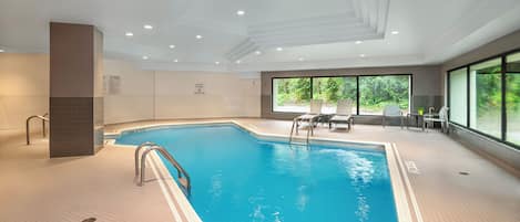 Une piscine intérieure avec un sol carrelé, un escalier en métal et une vue sur la verdure à l’extérieur, grâce à de grandes fenêtres.
