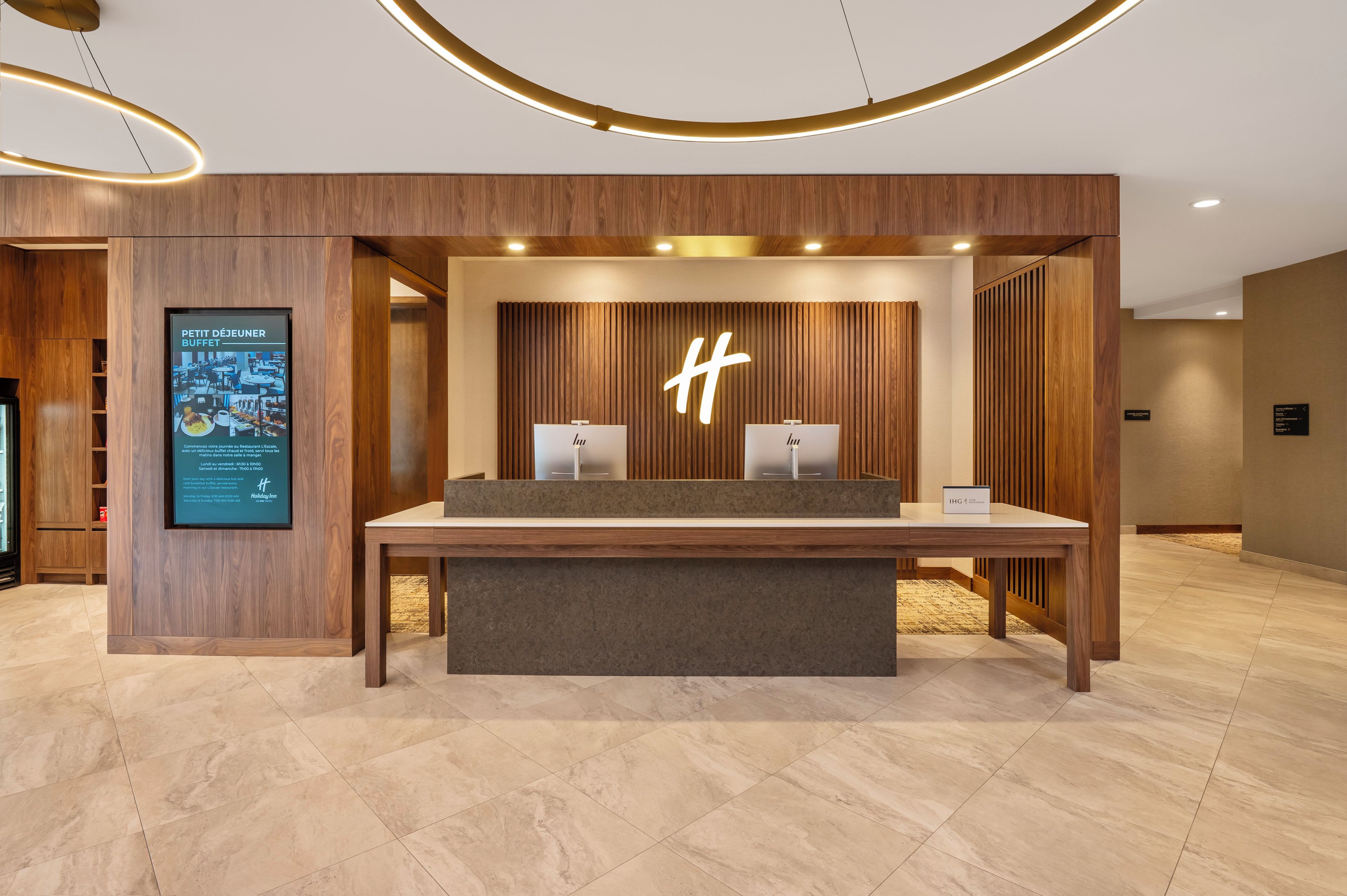 A reception desk with a wooden finish, a large illuminated 'H' sign, and a digital screen displaying hotel information.