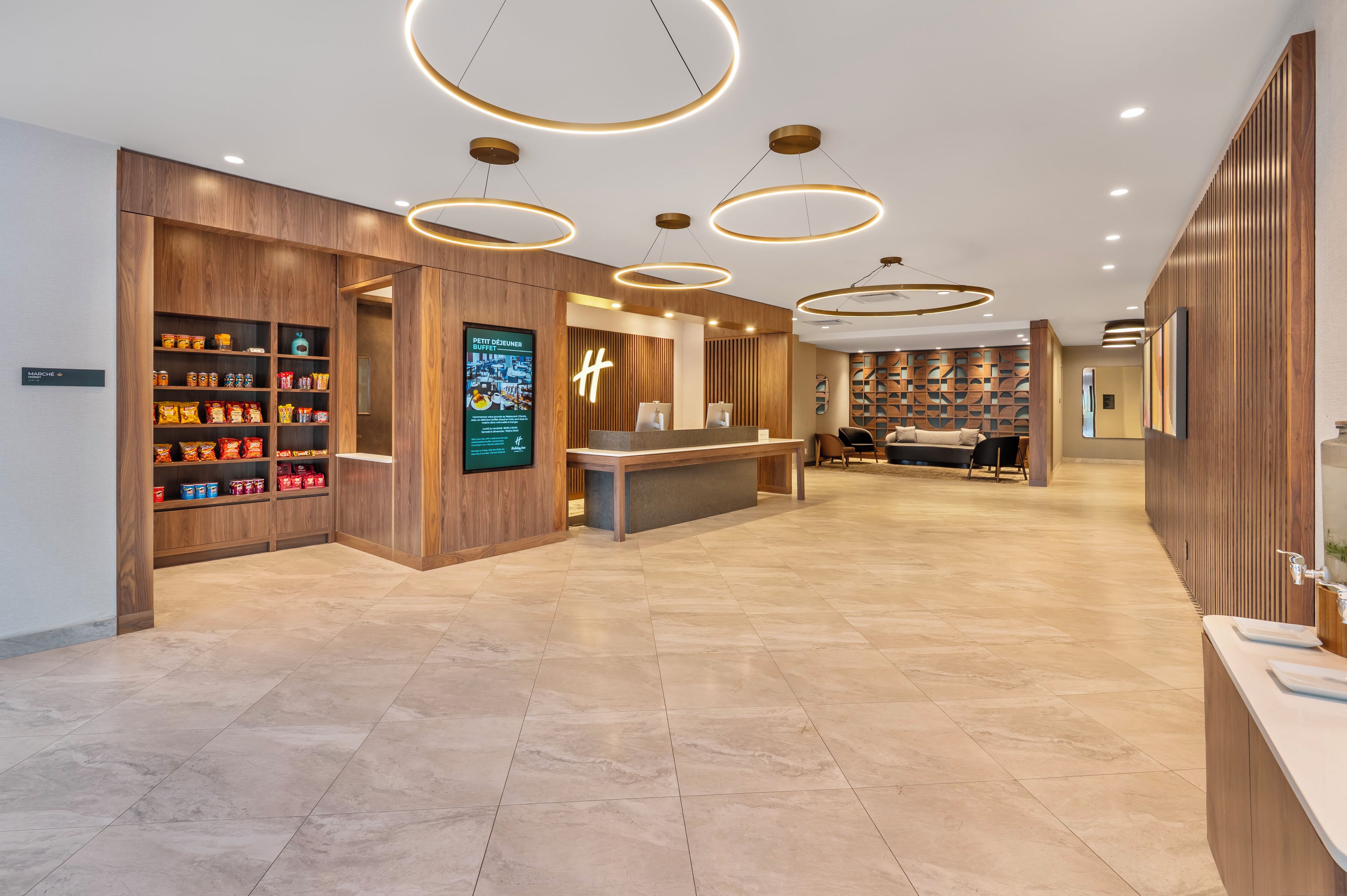 A hotel lobby with a reception desk, wooden shelving with snacks, and modern circular ceiling lights.