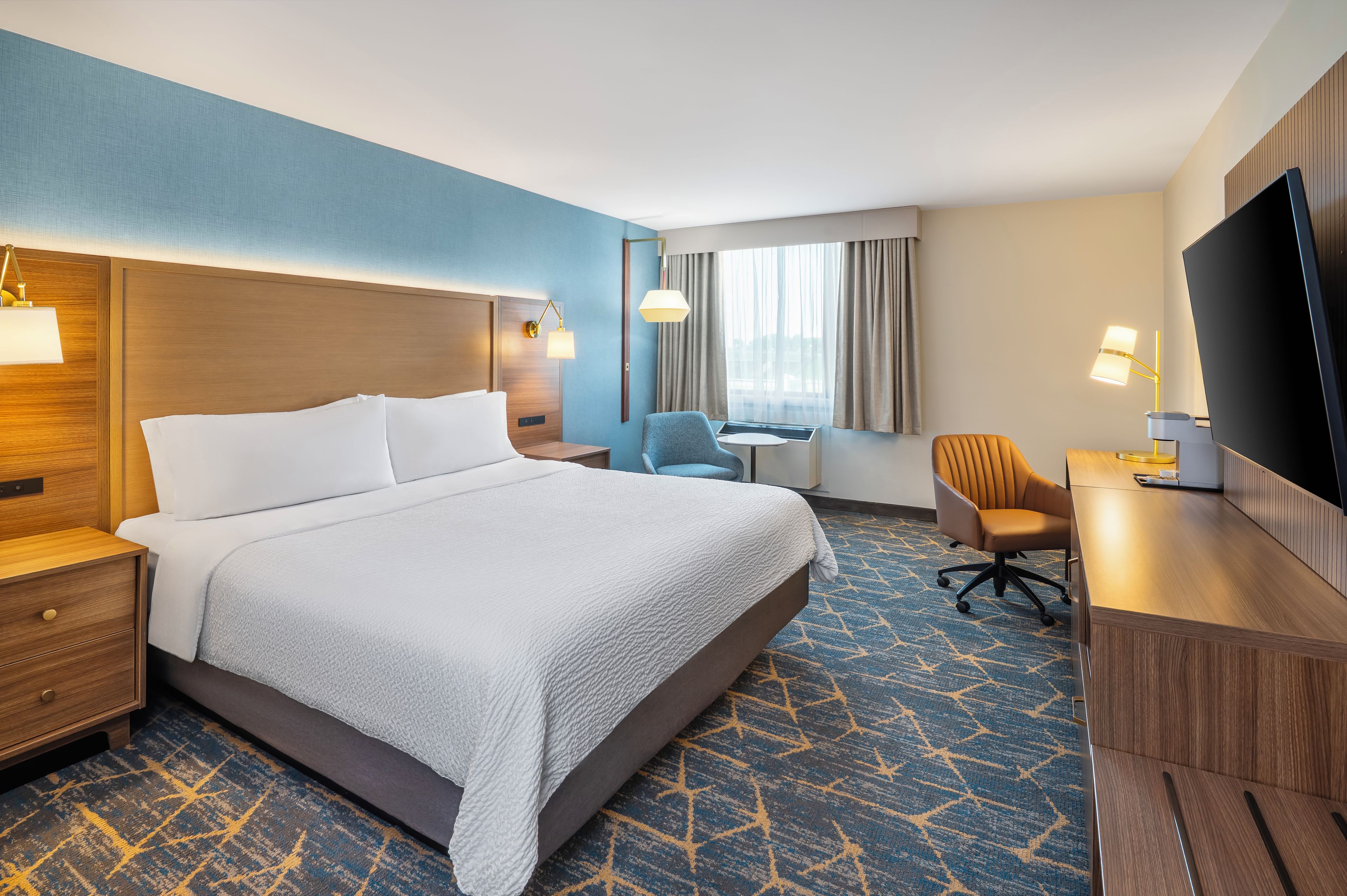 A hotel room with a large bed, a desk, a chair, a TV, and a window with curtains.