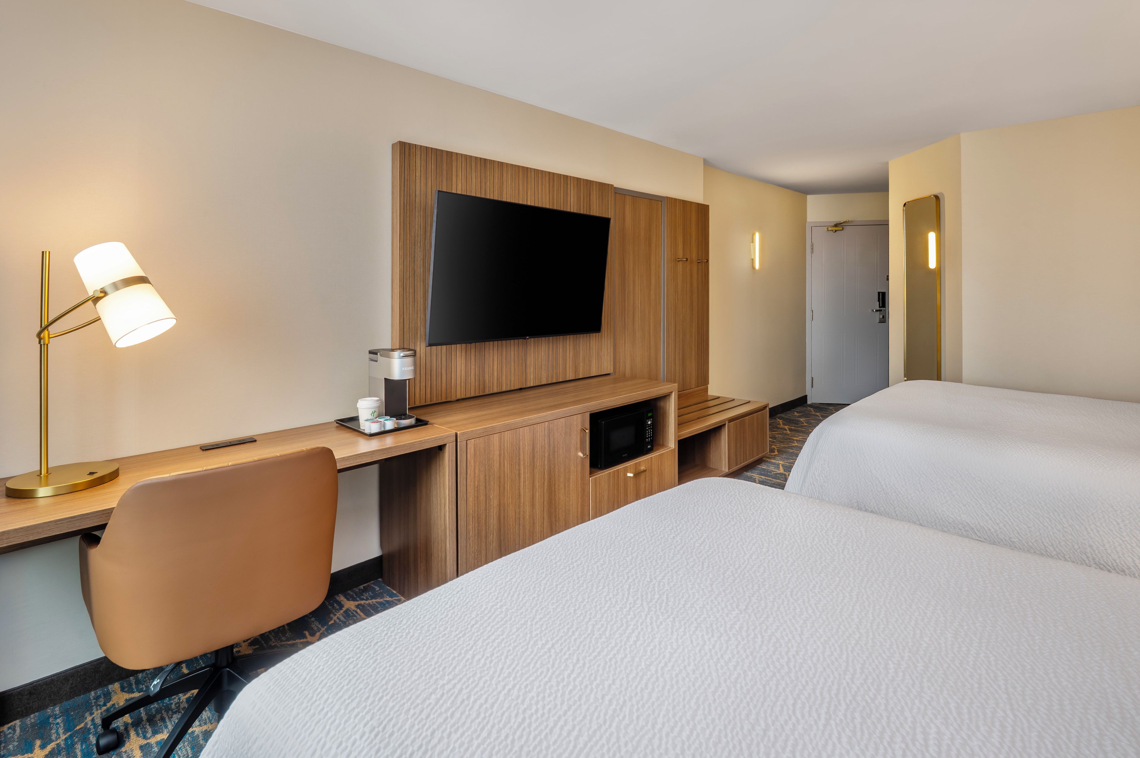 Ein Hotelzimmer mit Flachbildfernseher an einer Holzverkleidung, einem Schreibtisch mit Lampe, einer Mikrowelle und zwei Betten mit weißer Bettwäsche.