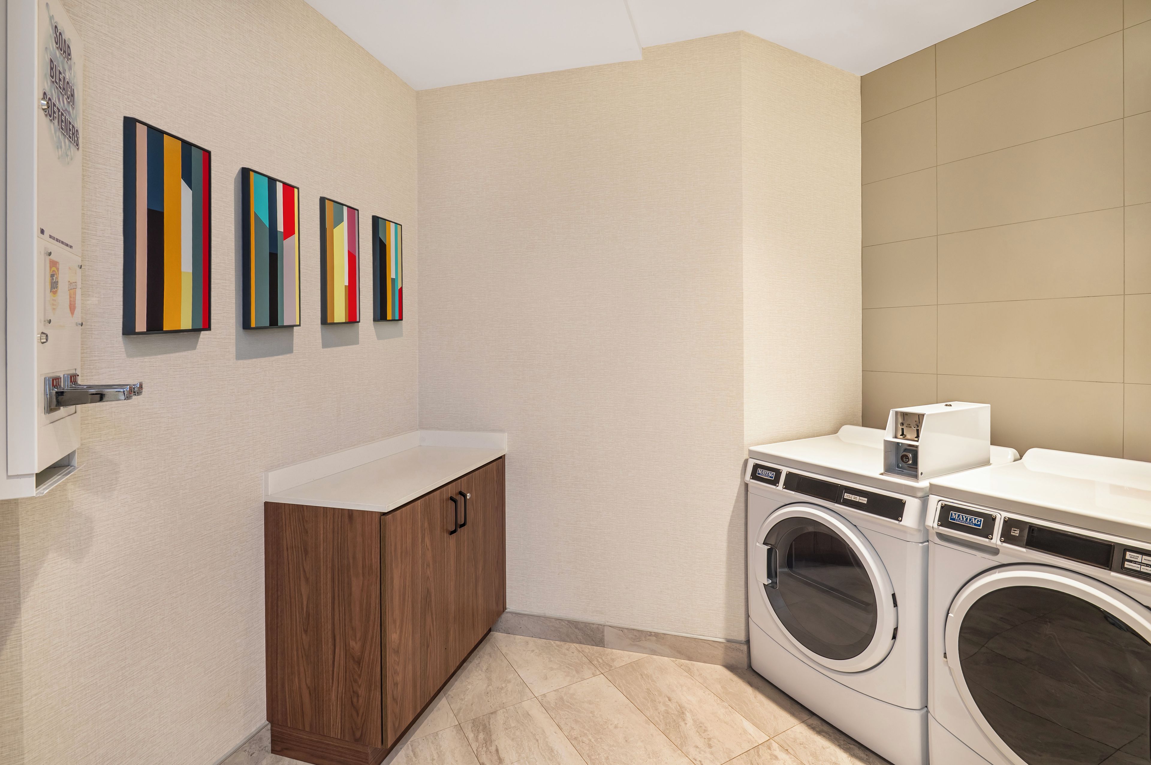 Un lavadero moderno con una lavadora, un gabinete de madera y tres cuadros abstractos y coloridos en la pared.