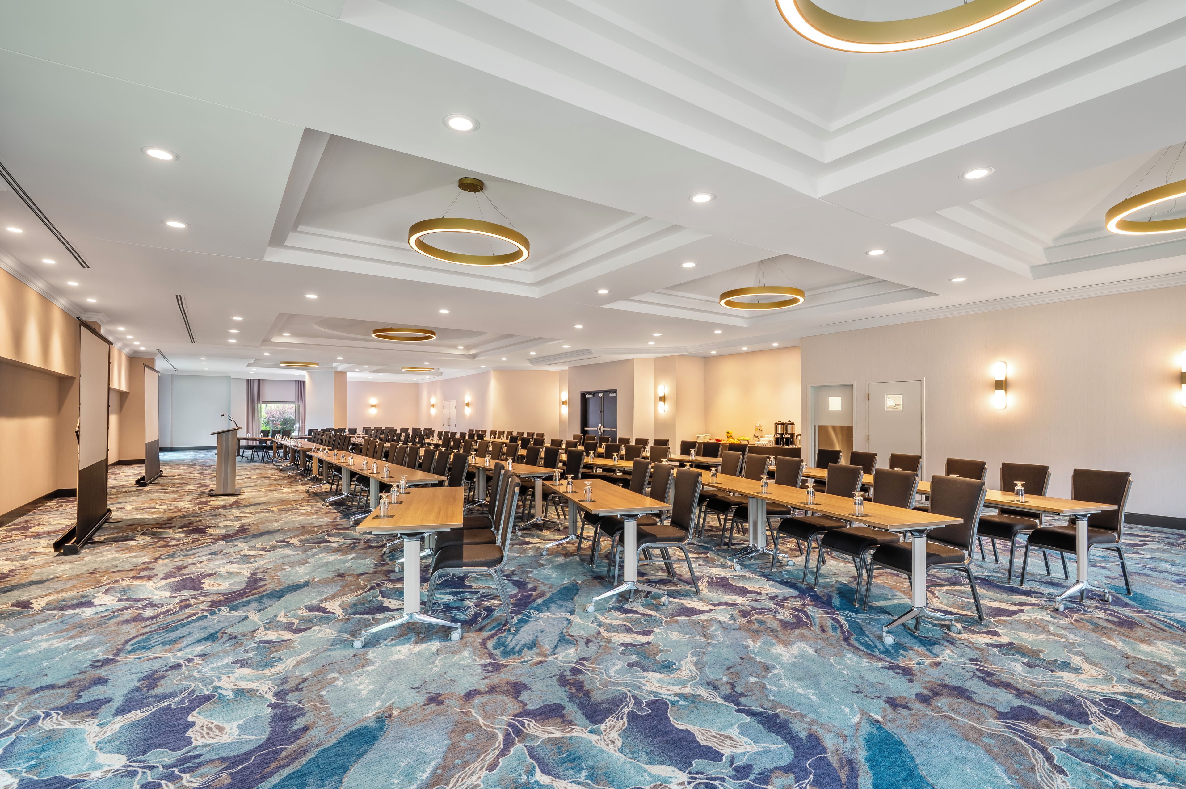 A conference room with rows of tables and chairs, a marble-patterned carpet, and recessed ceiling lighting.