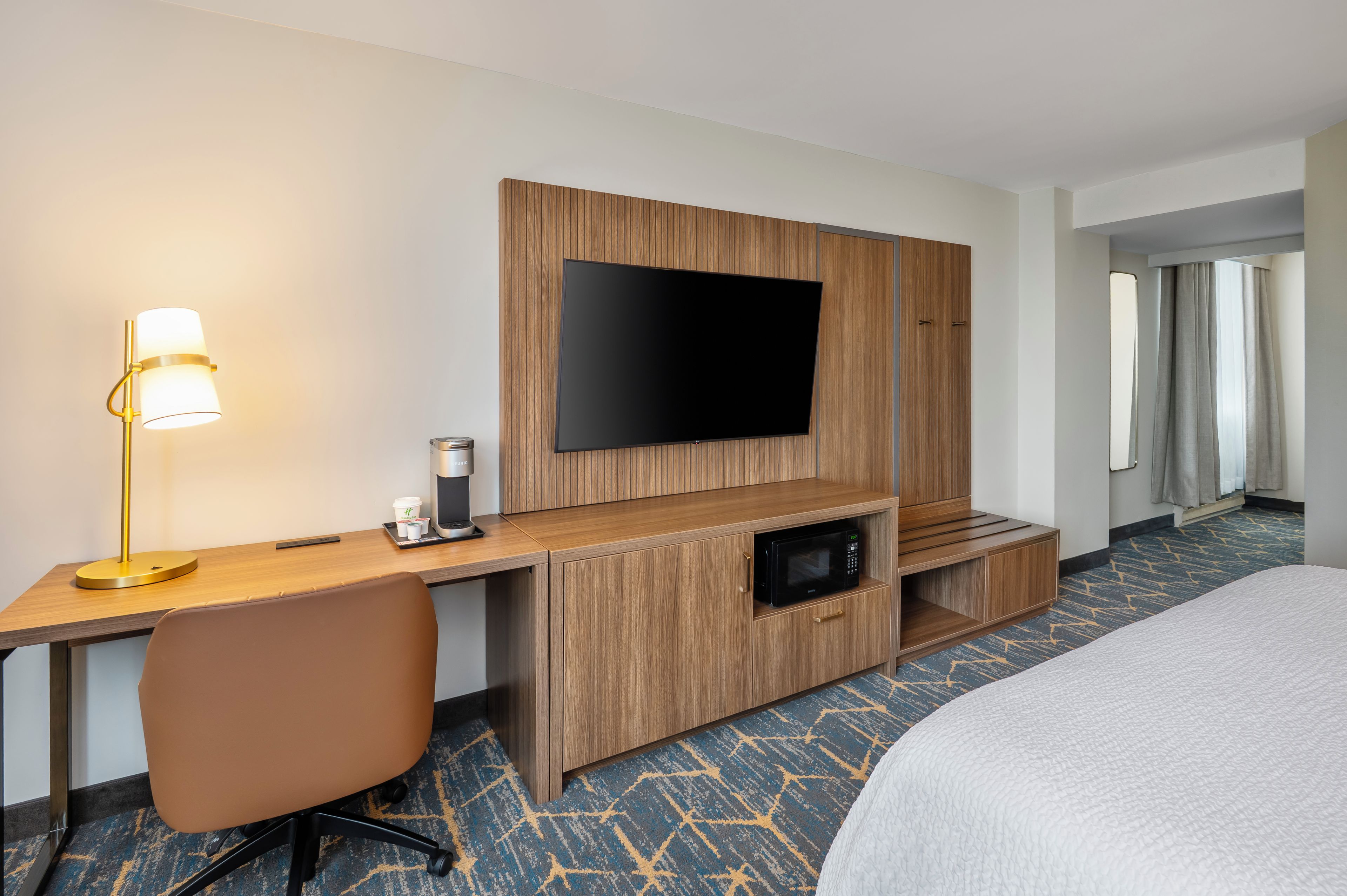 A hotel room with a wooden desk, a flat-screen TV, a coffee maker, a microwave, a bed, and a patterned carpet.