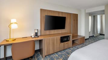 A hotel room with a wooden desk, a flat-screen TV, a coffee maker, a microwave, a bed, and a patterned carpet.