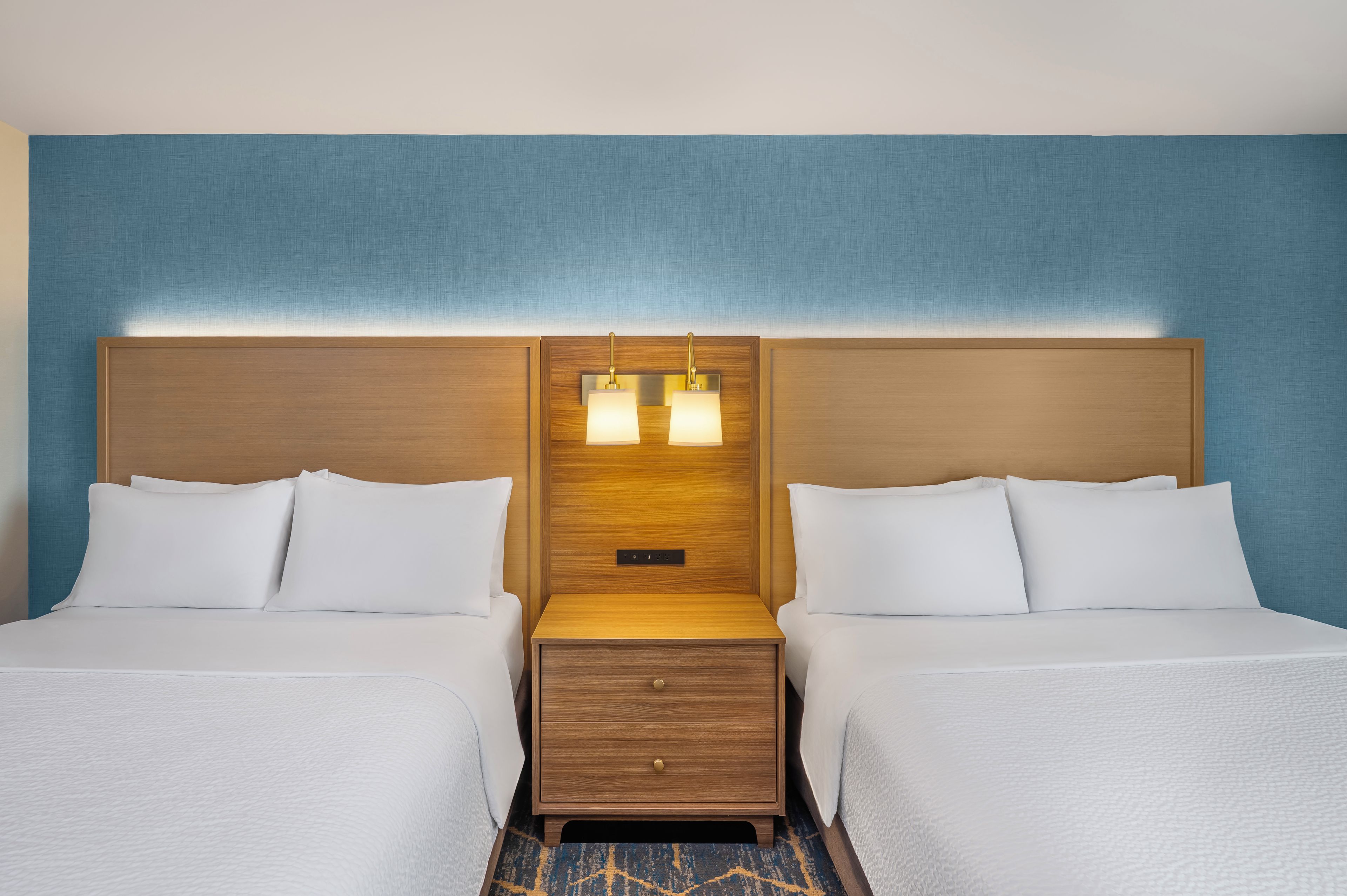 A hotel room with two beds, a wooden headboard, a nightstand with a lamp, and a blue wall.