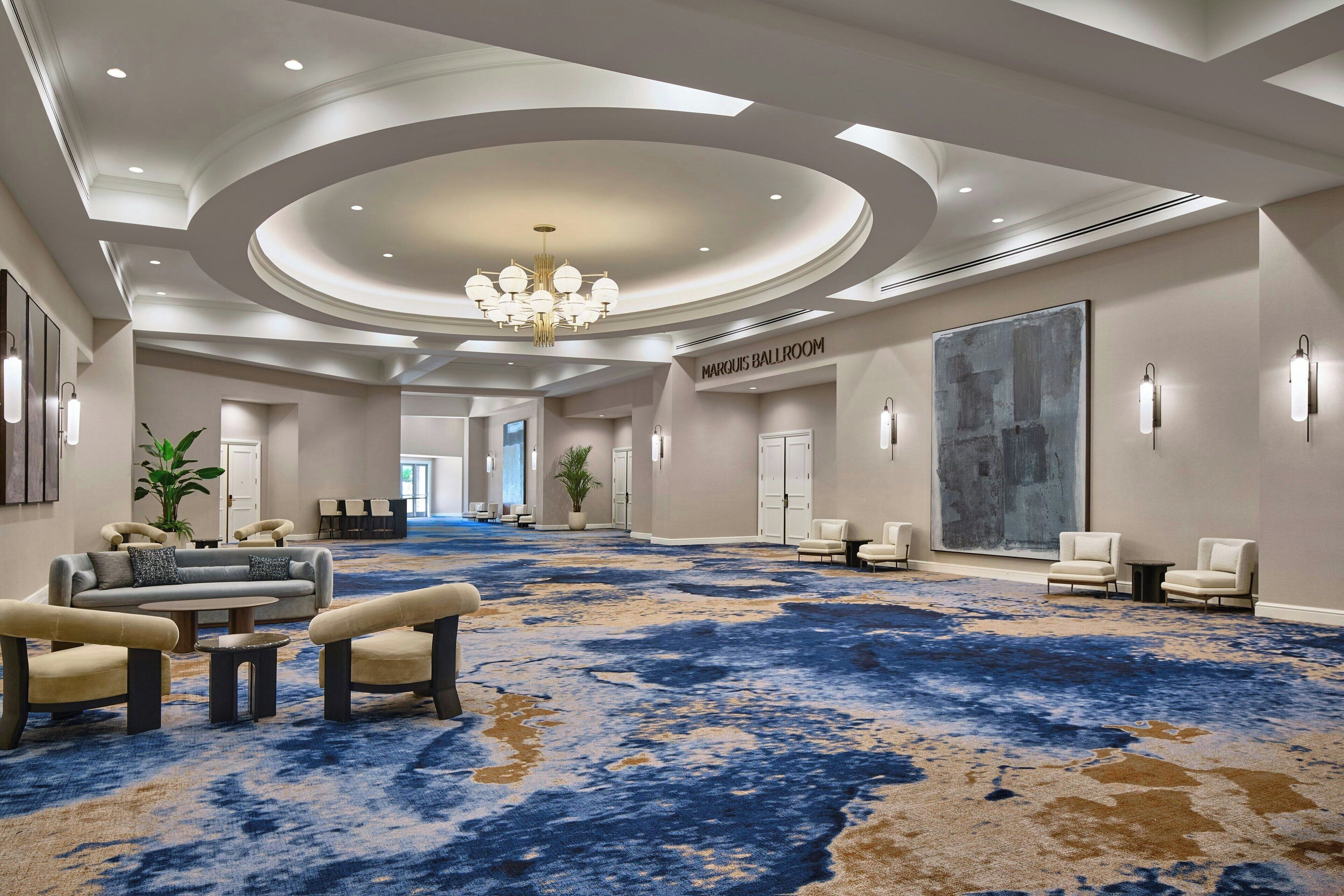 A spacious, well-lit hotel lobby with a blue and beige carpet, comfortable seating areas, and a large abstract painting on the wall.