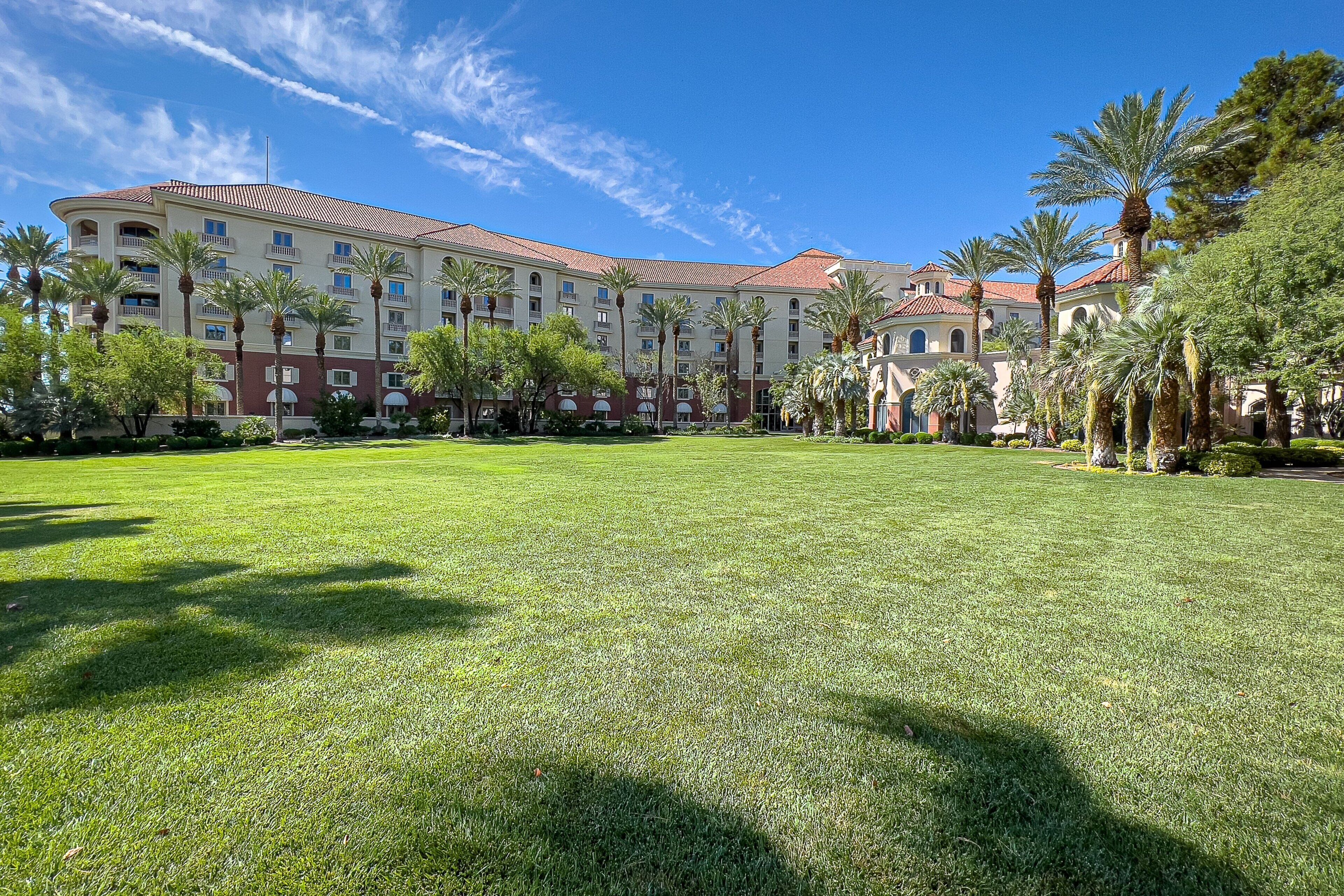 Un grande hotel con diversi piani, un cortile con palme e un cielo azzurro intenso.