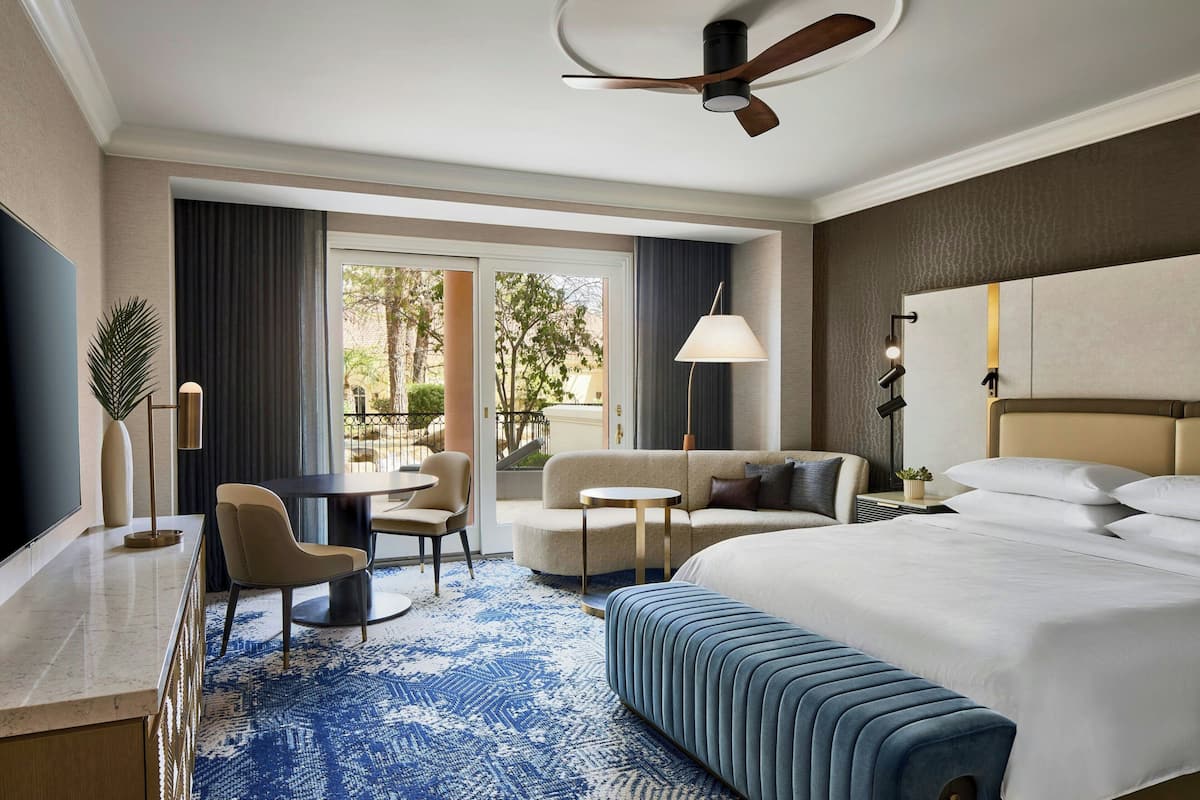 A hotel room with a large bed, a sofa, a desk, a chair, a small table, a lamp, and a ceiling fan.