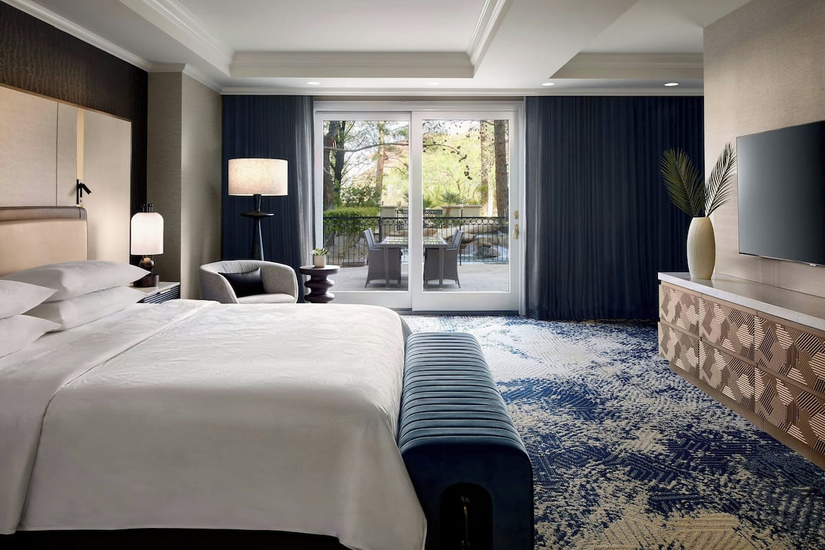 A hotel room with a large bed, a sofa, a TV, and a balcony with a view of trees.