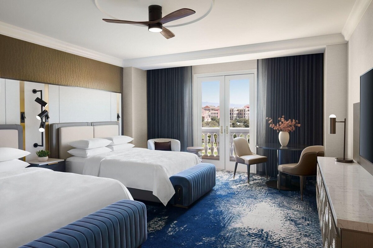 Habitación de hotel con dos camas, un balcón con vistas, un escritorio y un televisor.