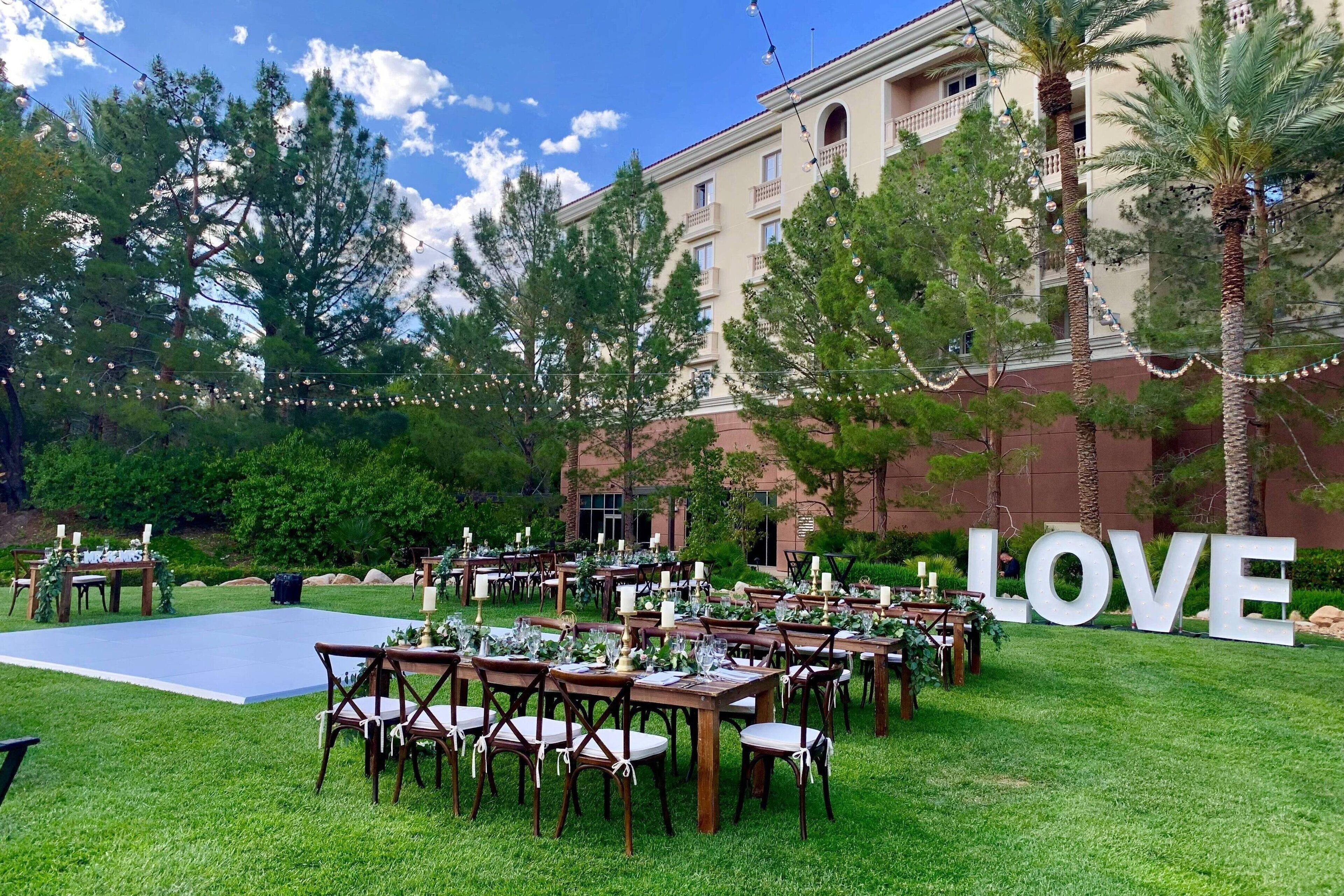 Une cérémonie de mariage en plein air, avec des tables, des chaises et une piste de danse, le tout entouré d’arbres et d’un bâtiment sur lequel est affiché un panneau avec le mot « AMOUR ».