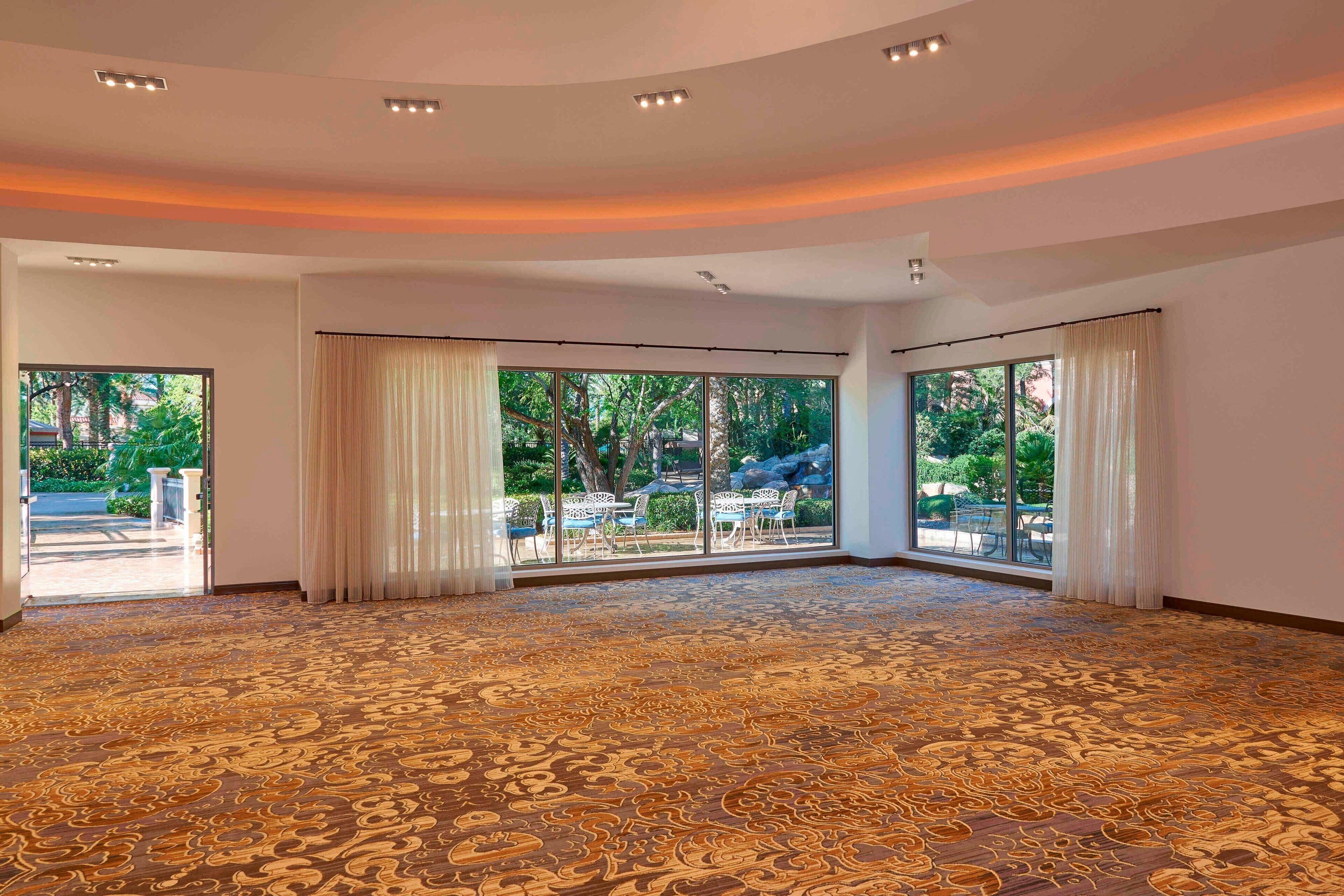 A spacious room with a patterned carpet, large windows with curtains, and a view of greenery outside.