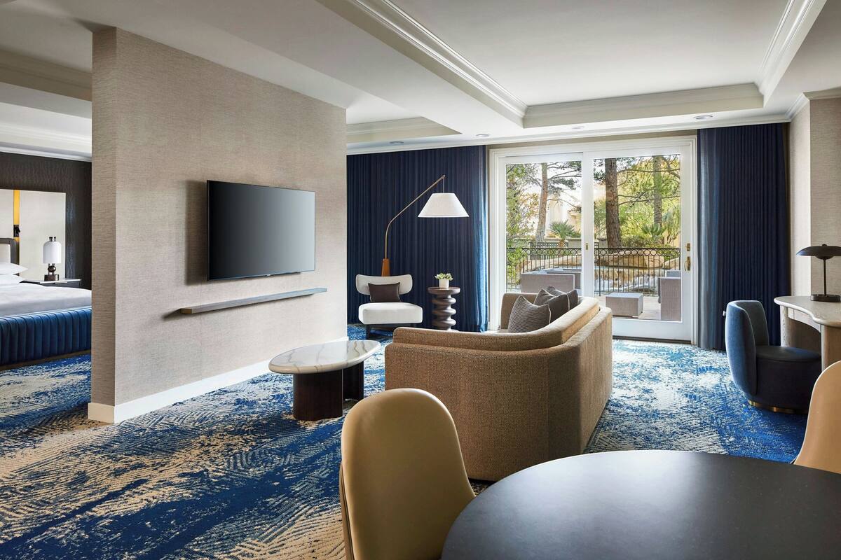 A modern hotel room with a large flat-screen TV mounted on the wall, a comfortable sofa, a dining table, and a balcony with a view of the outdoors.