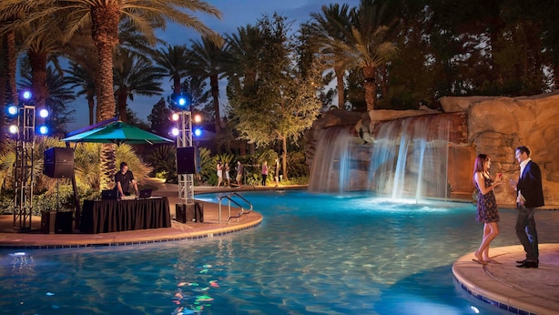 A pool area with a waterfall feature, palm trees, and a DJ setup.