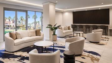 Un moderno hall de hotel con una amplia zona de estar, un bar y grandes ventanales que ofrecen vistas a palmeras.