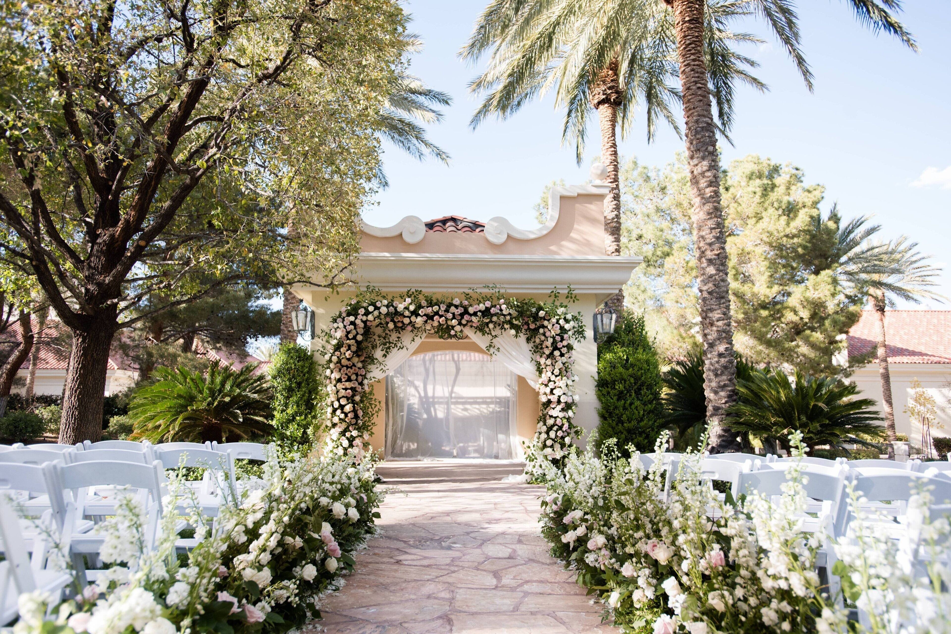 An outdoor wedding aisle with white chairs and a floral archway.