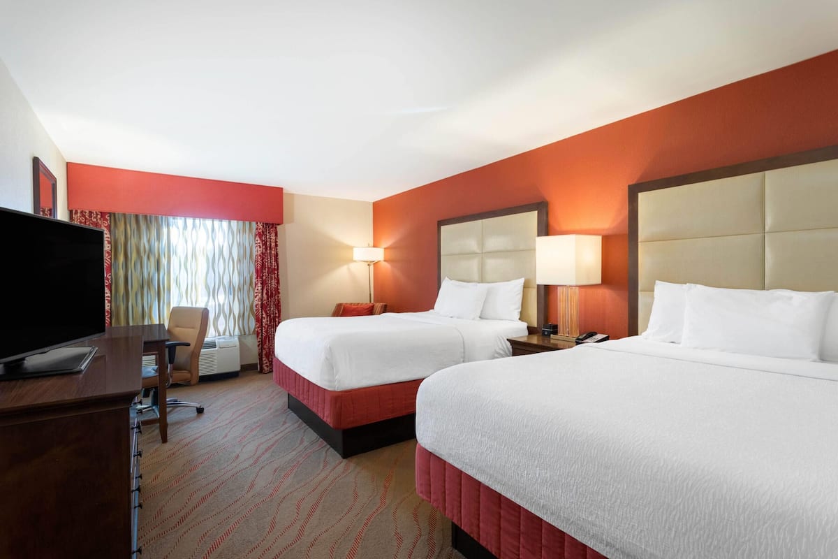 A hotel room with two beds, a desk with a flat-screen TV, a chair, and a lamp.