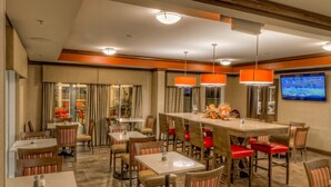 A hotel dining area with multiple tables and chairs, a central bar area, and a television mounted on the wall.
