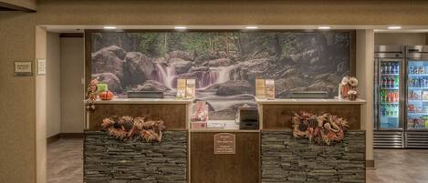Mostrador de recepción con un mural de cascada, un cartel que indica cien por ciento libre de humo y un refrigerador lleno de bebidas.