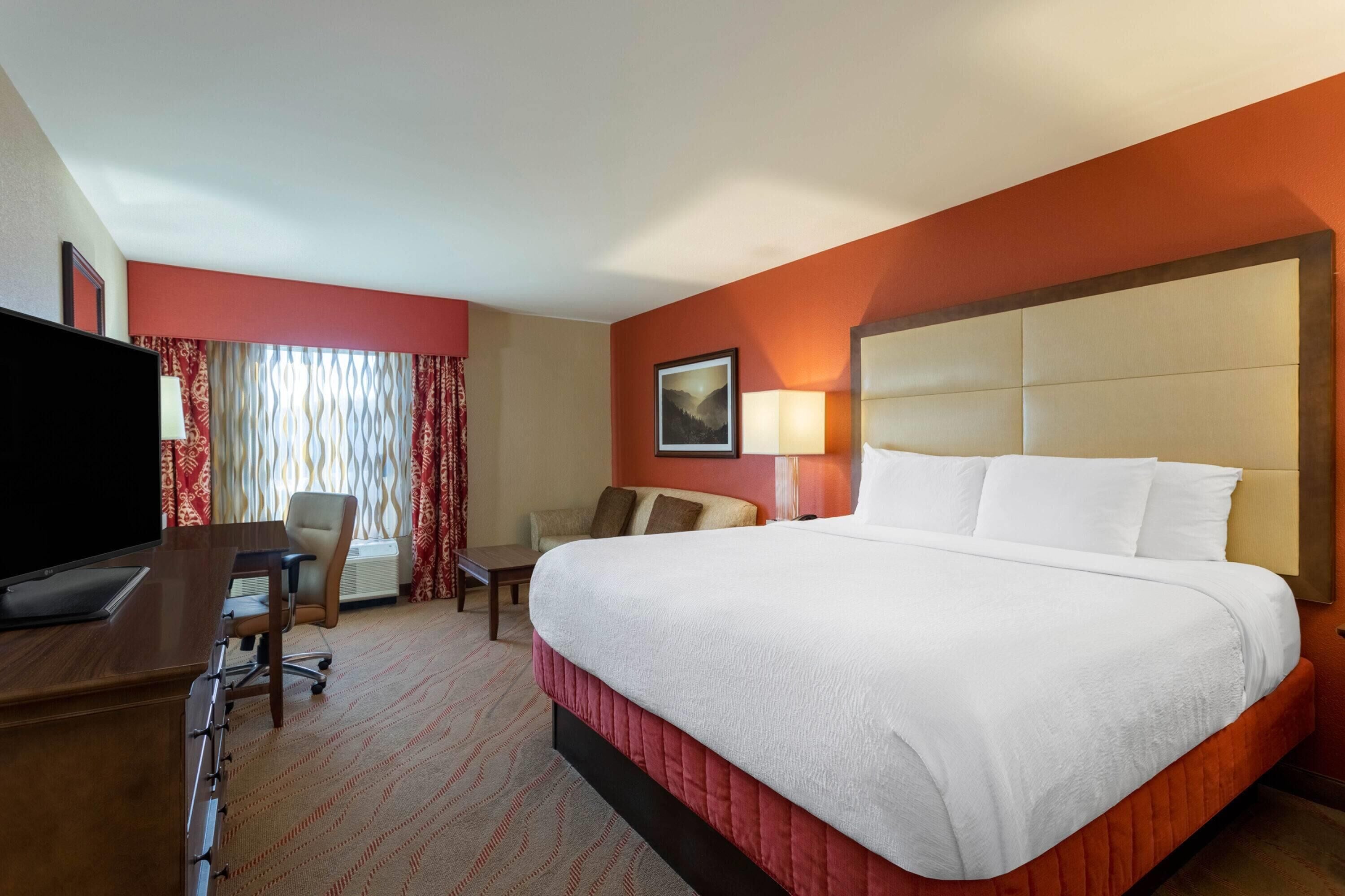 Quarto de hotel com uma cama grande, uma escrivaninha com televisão, uma cadeira e uma janela com cortinas.