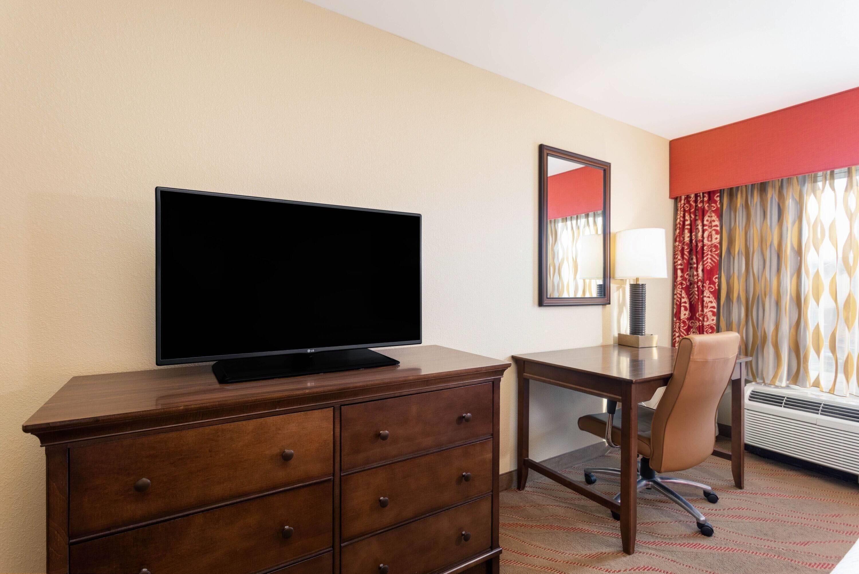 Habitación de hotel con cómoda de madera, televisor de pantalla plana, escritorio con silla, espejo y ventana con cortinas estampadas.