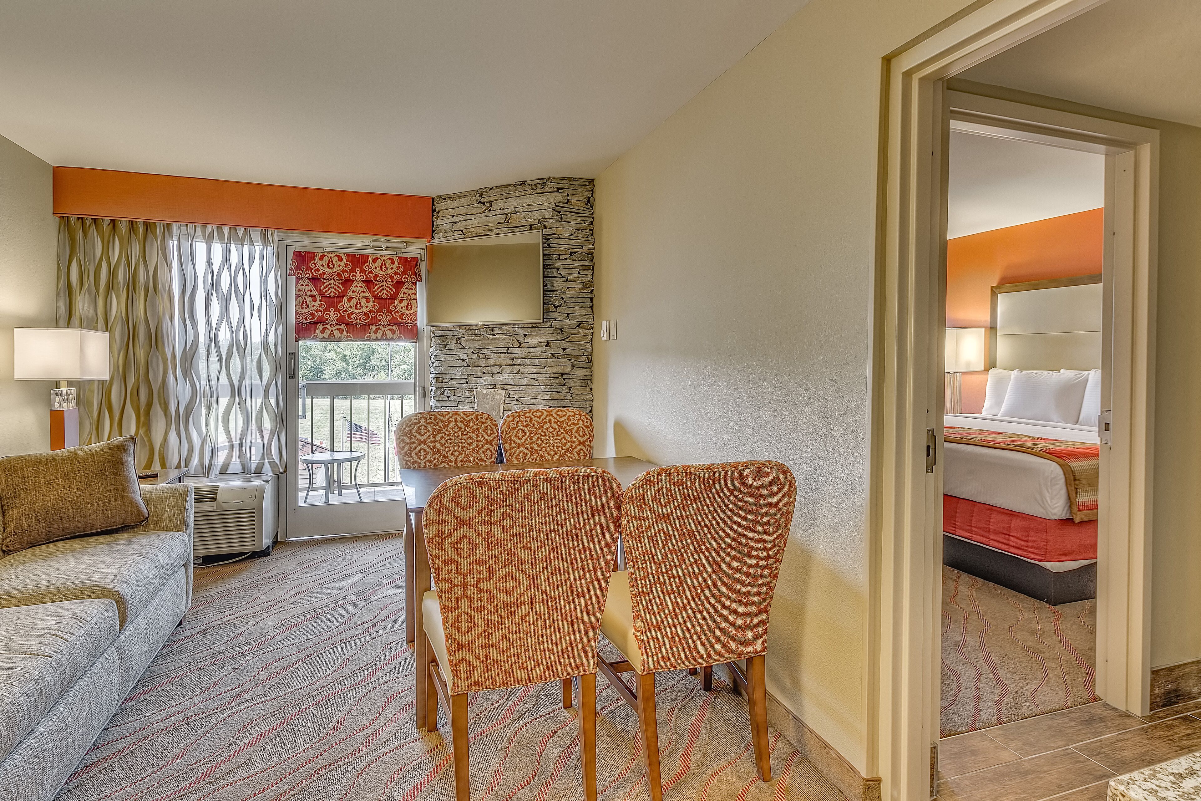 A hotel room with a dining area, a sofa, patterned chairs, a stone accent wall, and a bed with a red bedspread.