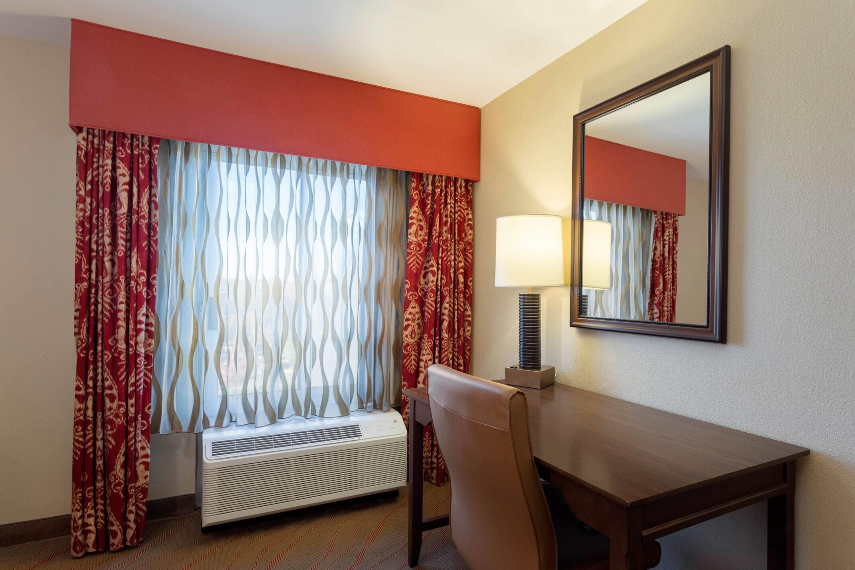 Een hotelkamer met een bed, een bureau met lamp, een raam met gordijnen met patroon en een spiegel aan de muur.
