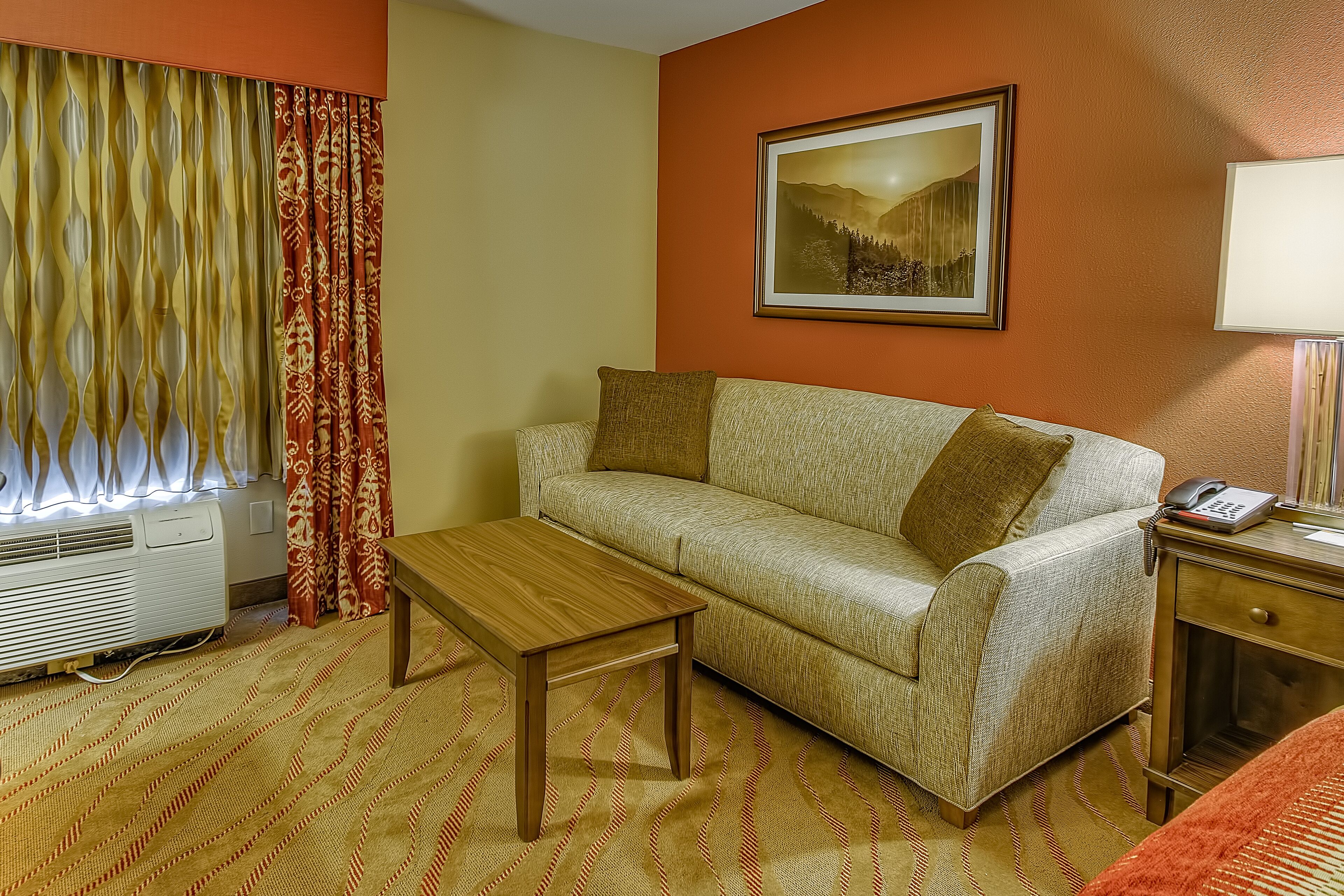 A hotel room with a sofa, a wooden table, a lamp, and a painting on the wall.