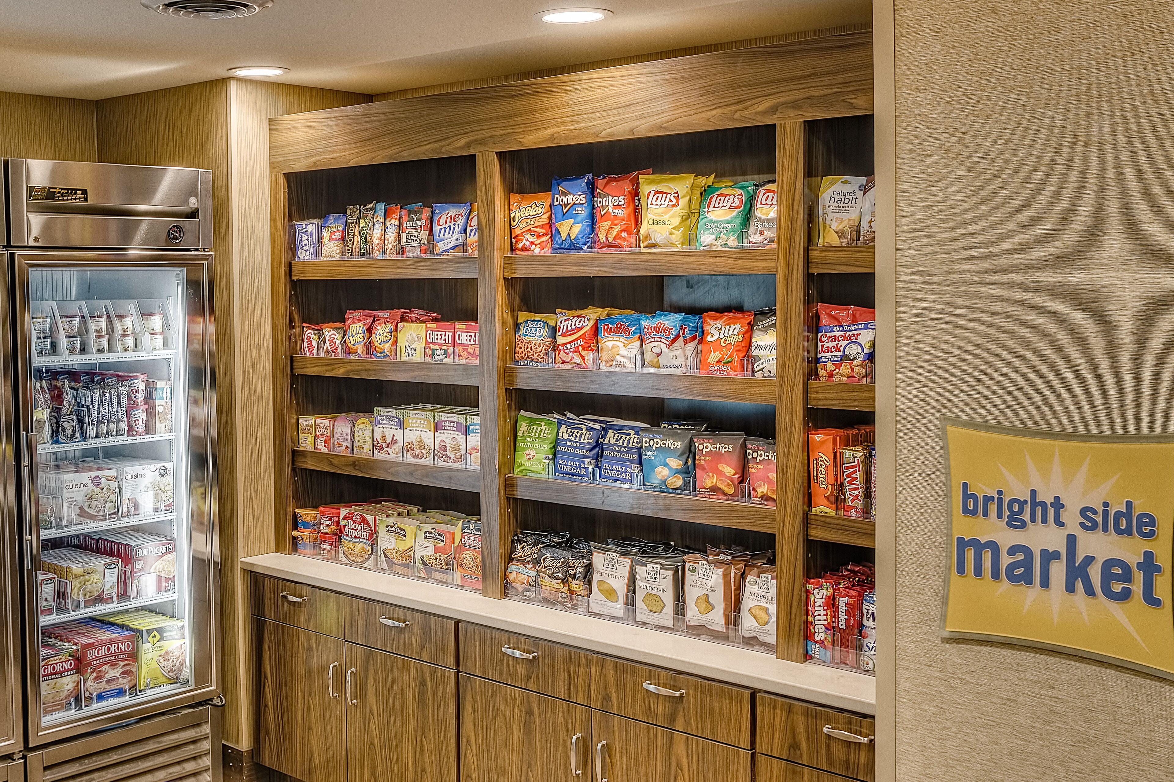 Un expositor de productos de alimentación con diferentes aperitivos y cereales, un refrigerador con bebidas, y un letrero que dice 'bright side market'.