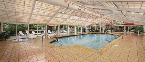 Una piscina interior con suelo de baldosas, techo con tragaluces y sillones de descanso al exterior.