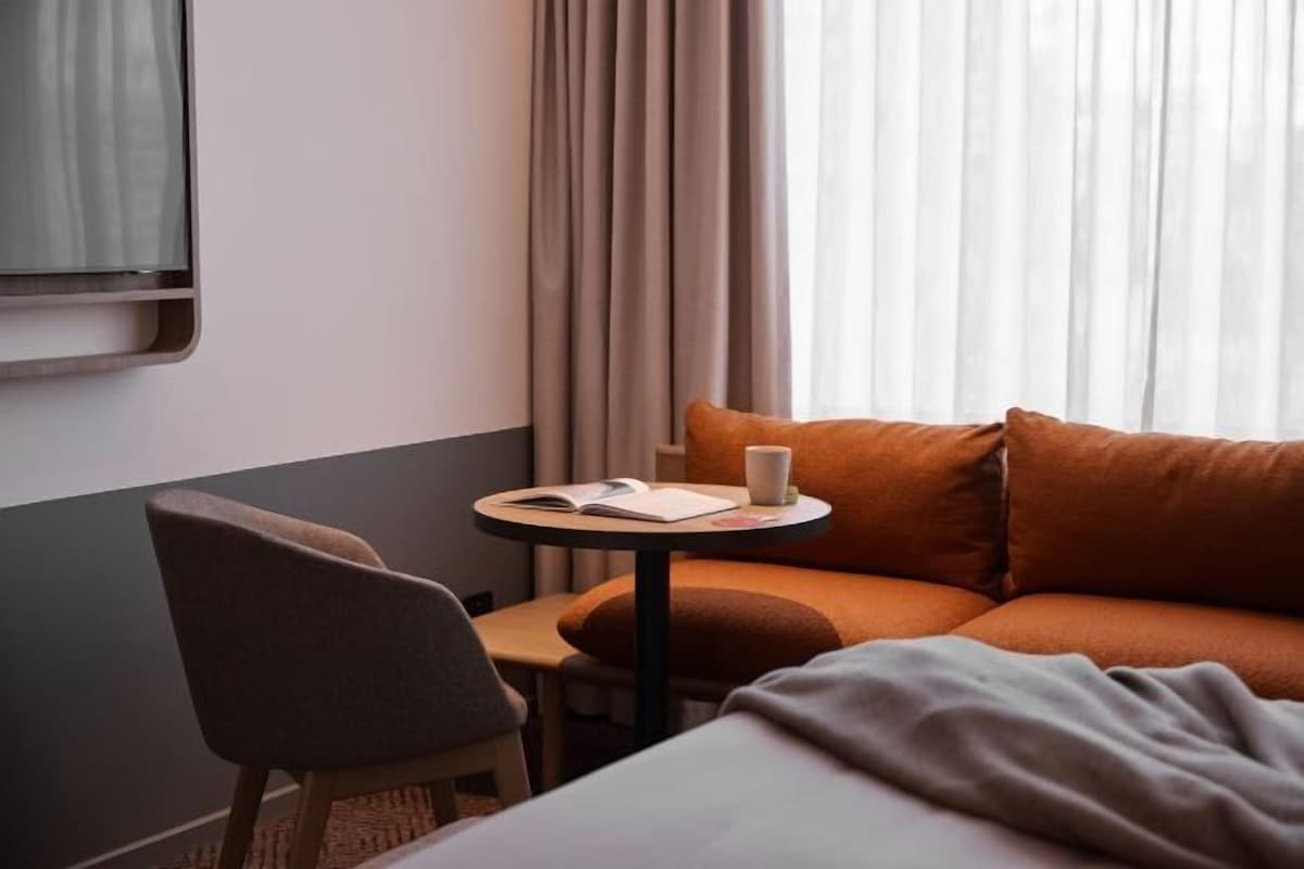A hotel room with a bed, a small round table with a book and a cup, a chair, and a window with curtains.