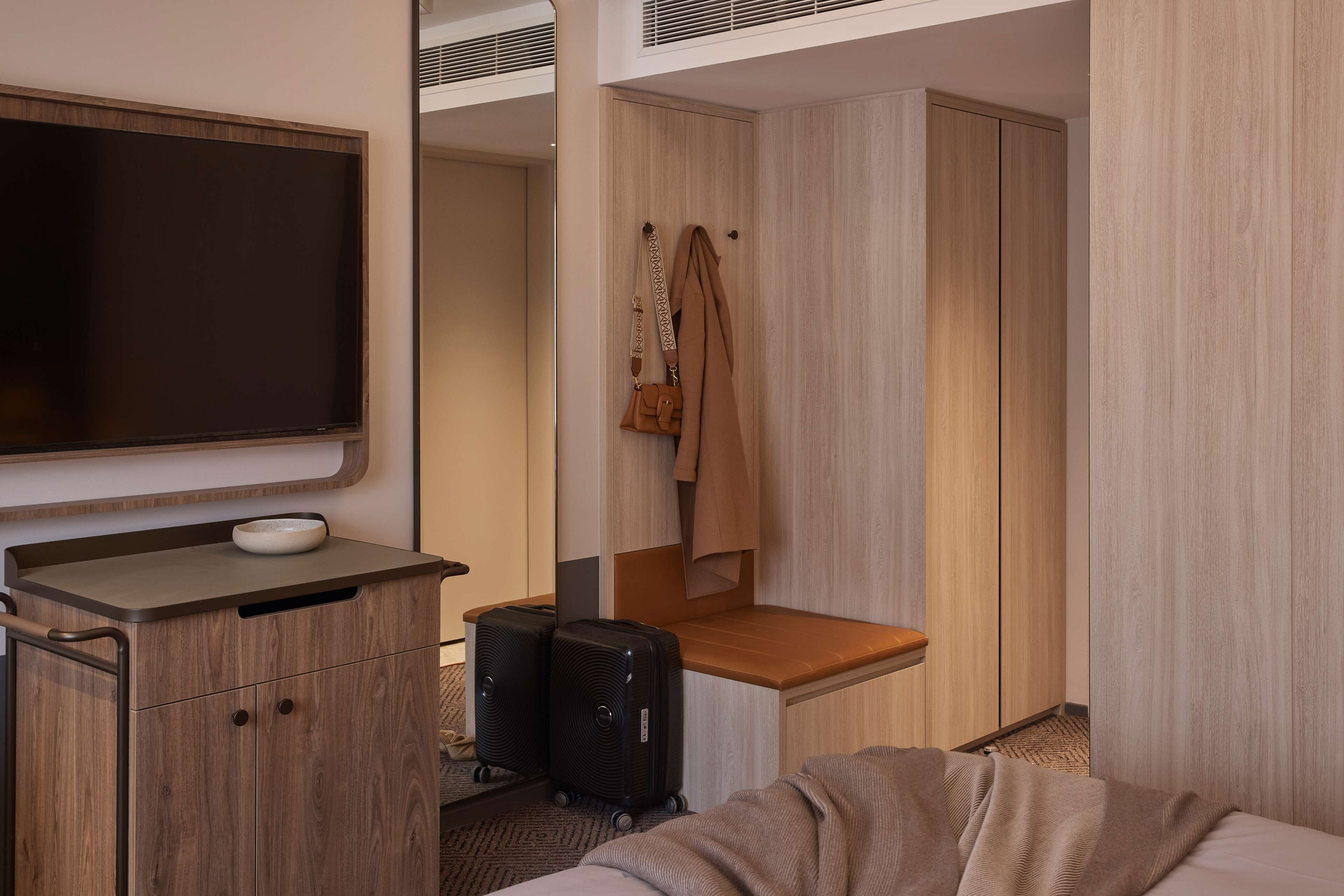 A hotel room with a flat-screen TV, wooden wardrobe, and a seating area with a bench.