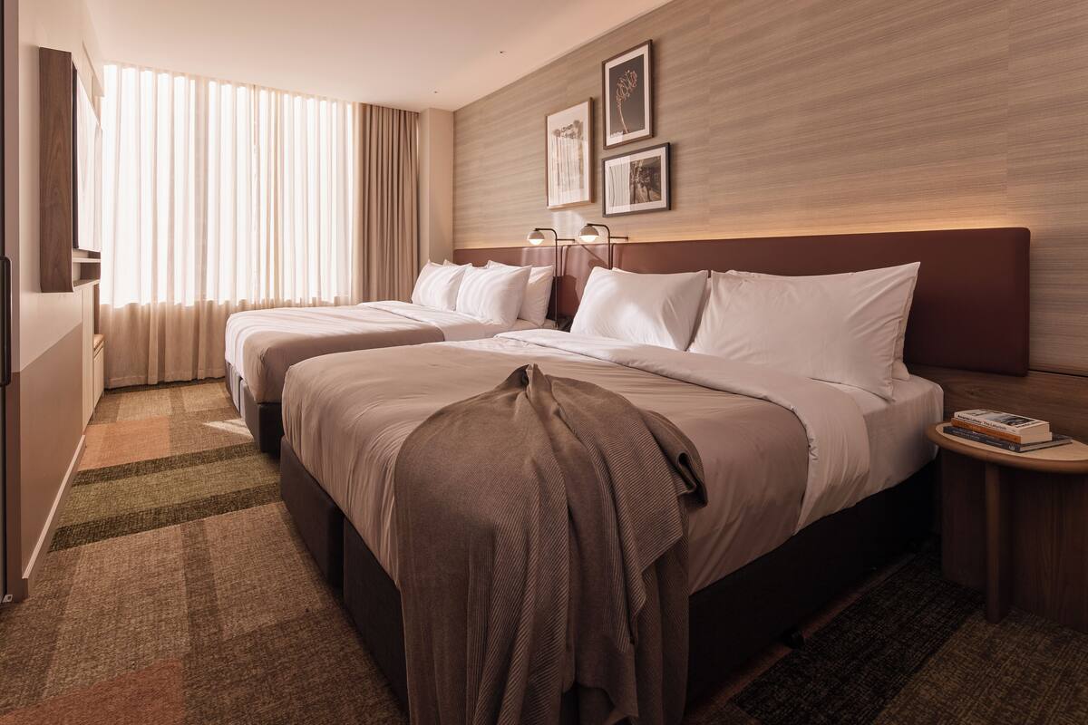 Ein Hotelzimmer mit zwei Betten, einem großen Fenster mit Vorhängen, einem Wandfernseher und gerahmten Bildern.