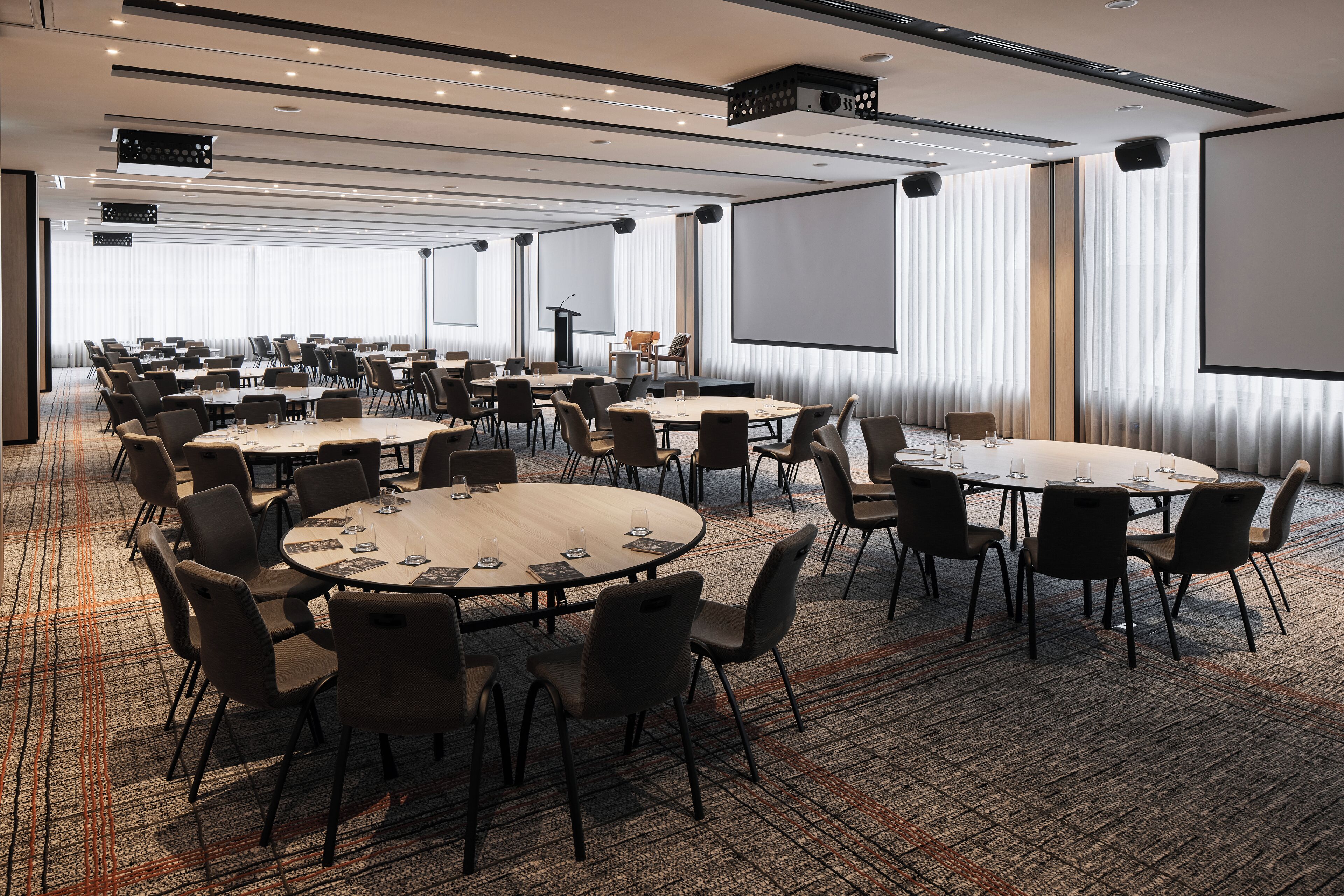 A conference room with multiple tables and chairs, large windows with curtains, a projector screen, and a podium.