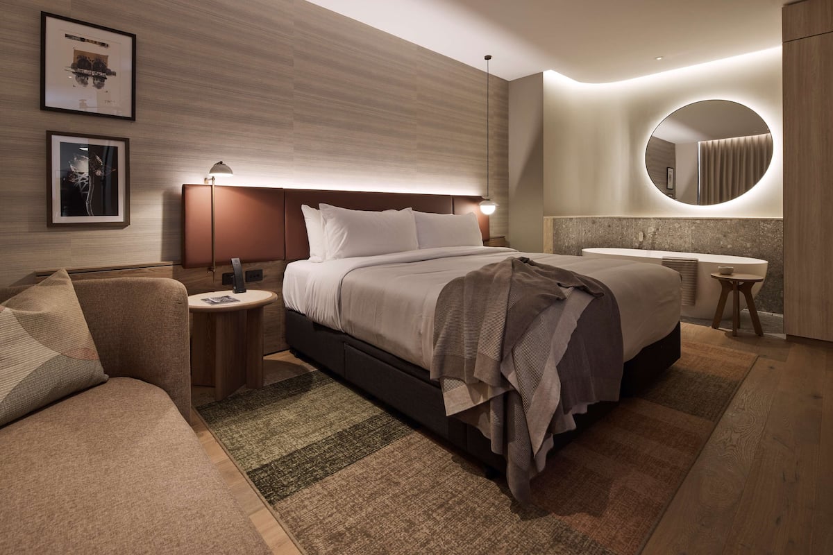 Ein modernes Hotelzimmer mit einem großen Bett, einem Nachttisch, einem runden Spiegel und einem kleinen Hocker.