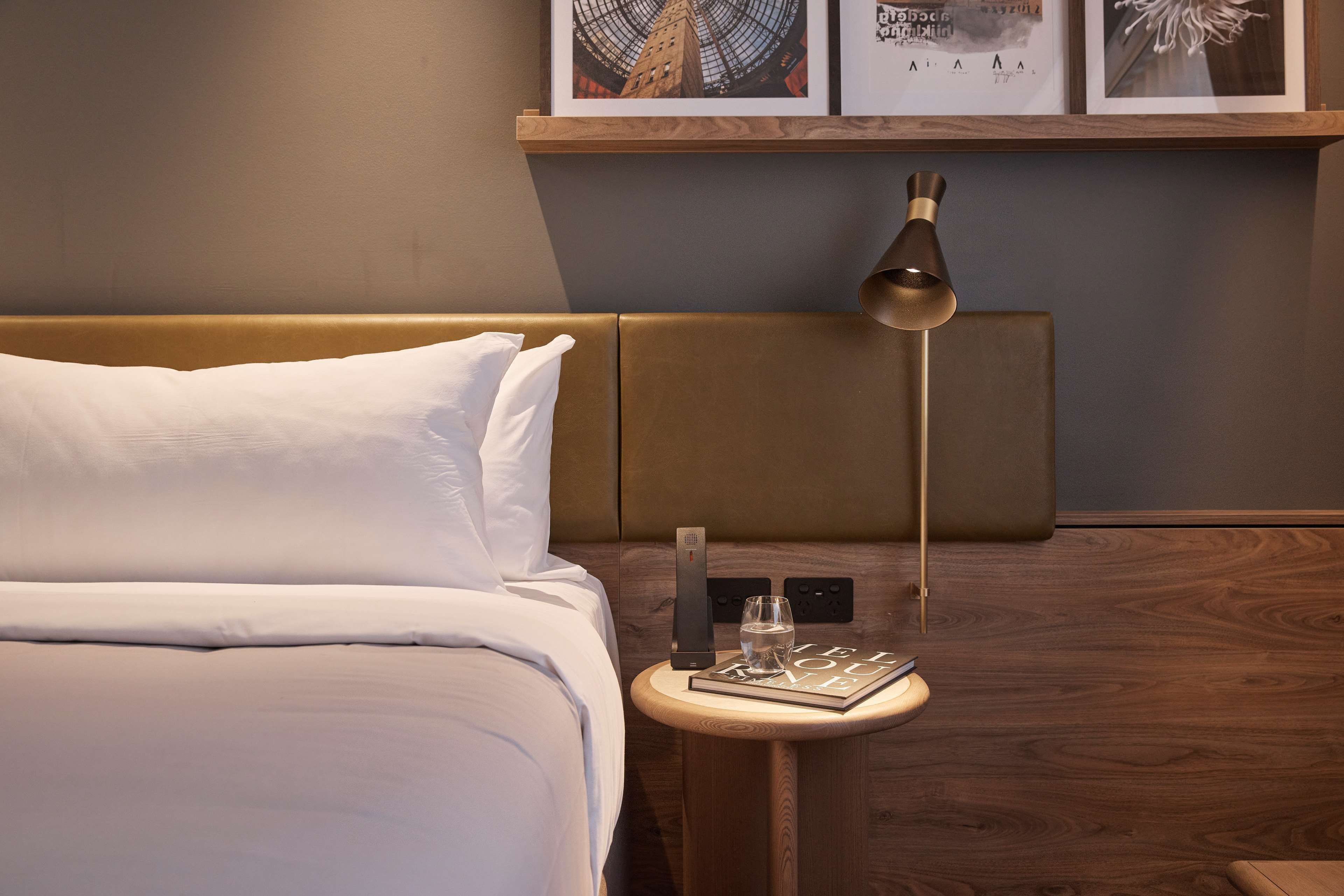 A neatly made bed with white linens, a bedside table with a lamp, a glass of water, and a book titled "Hello World".