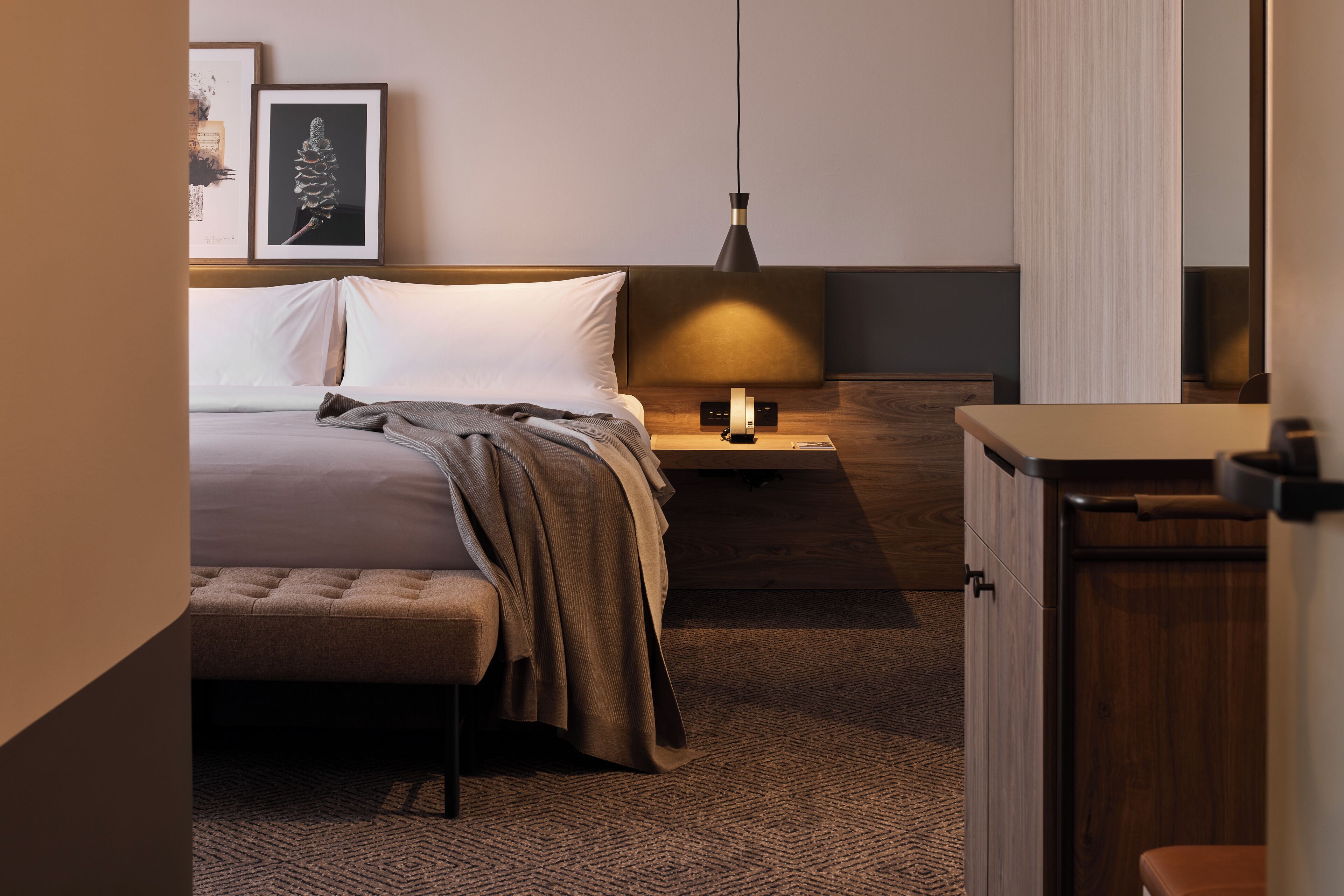 Una habitación de hotel ordenada con cama, mesita de noche, lámpara y un armario pequeño.