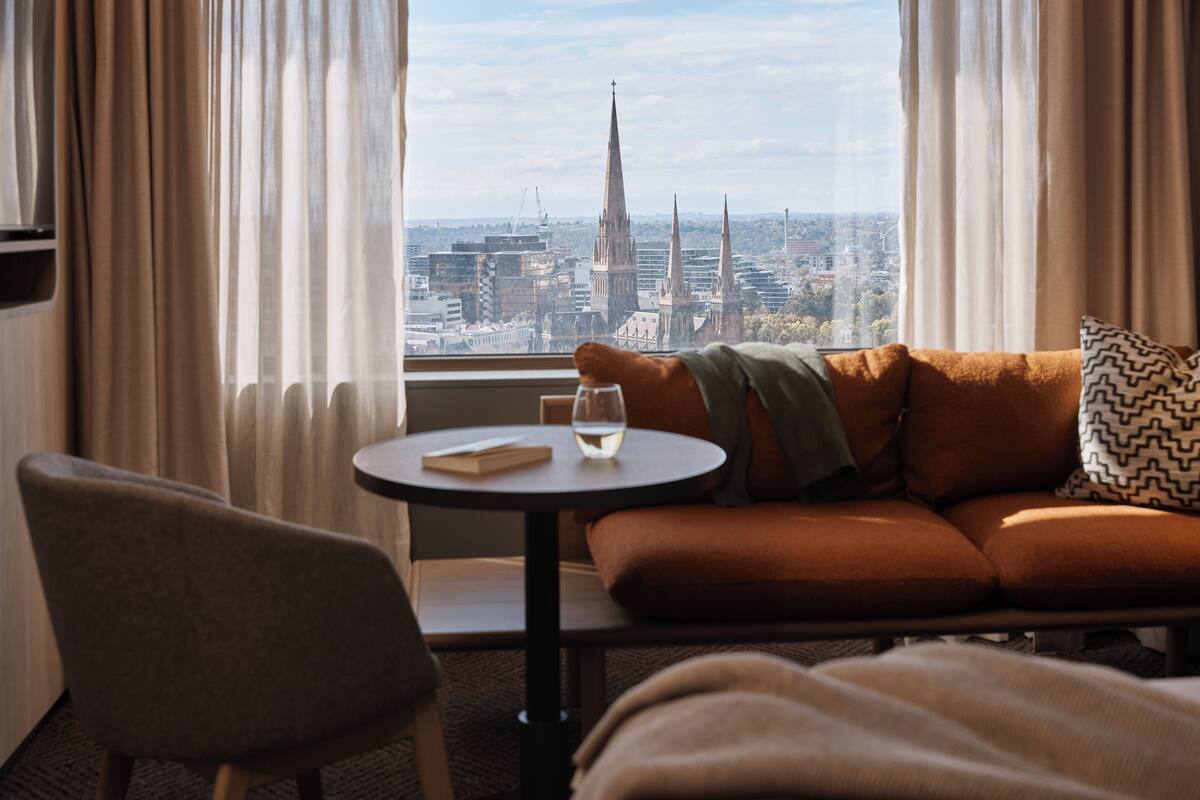 Ein Hotelzimmer mit einer Couch, einem kleinen Tisch mit einem Glas Wasser und einem Buch, und Blick auf eine Skyline mit hohen Gebäuden.